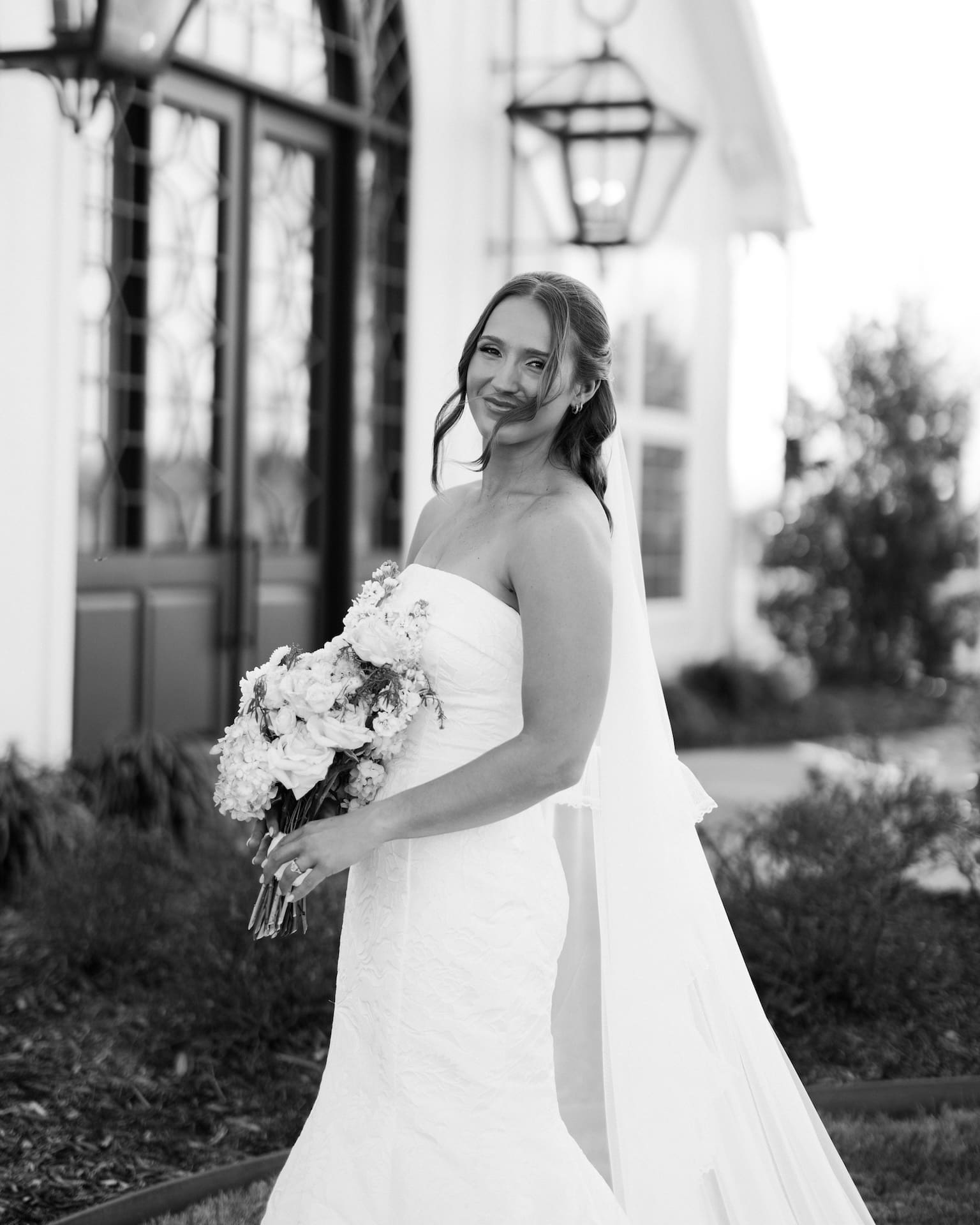 Black and white photo of a bride in a strapless lace mermaid gown glancing back with a smile in front of iron-paned chapel doors and glowing lanterns
