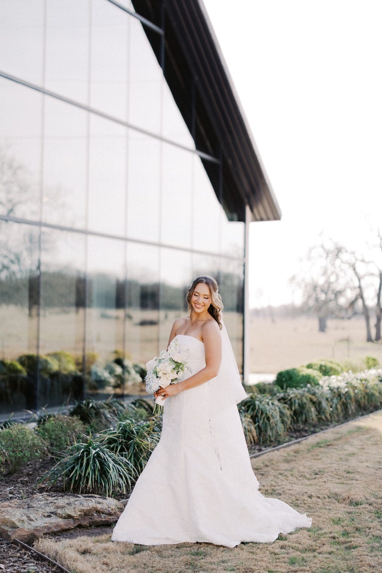 A bride stands in front of a glass barn facade at sunset, smiling down at her all-white bouquet, the countryside reflected behind her