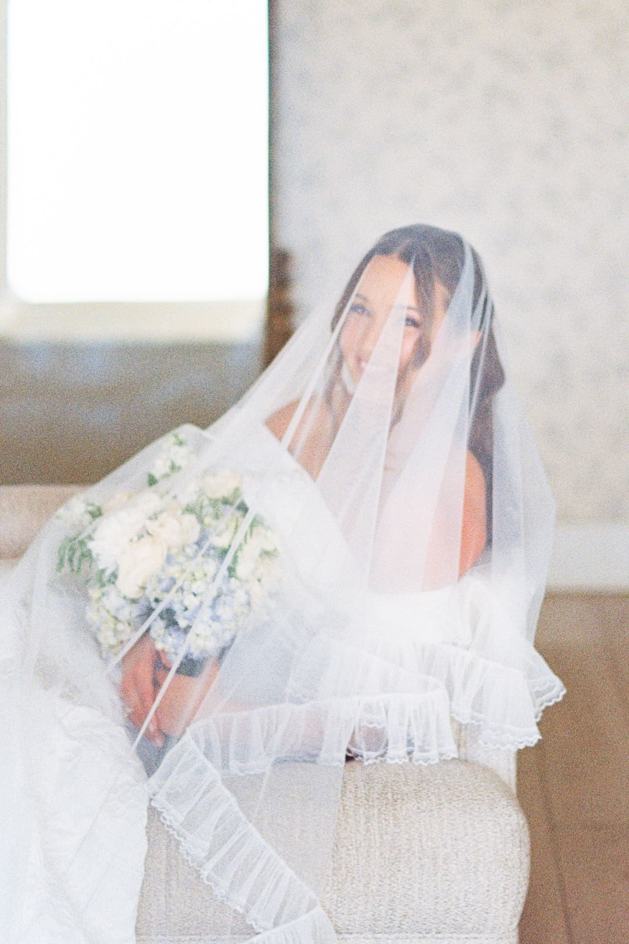 A bride seated and draped entirely in her veil peers out with a soft smile, bouquet of white and blue flowers resting in her lap in a warmly lit bridal suite