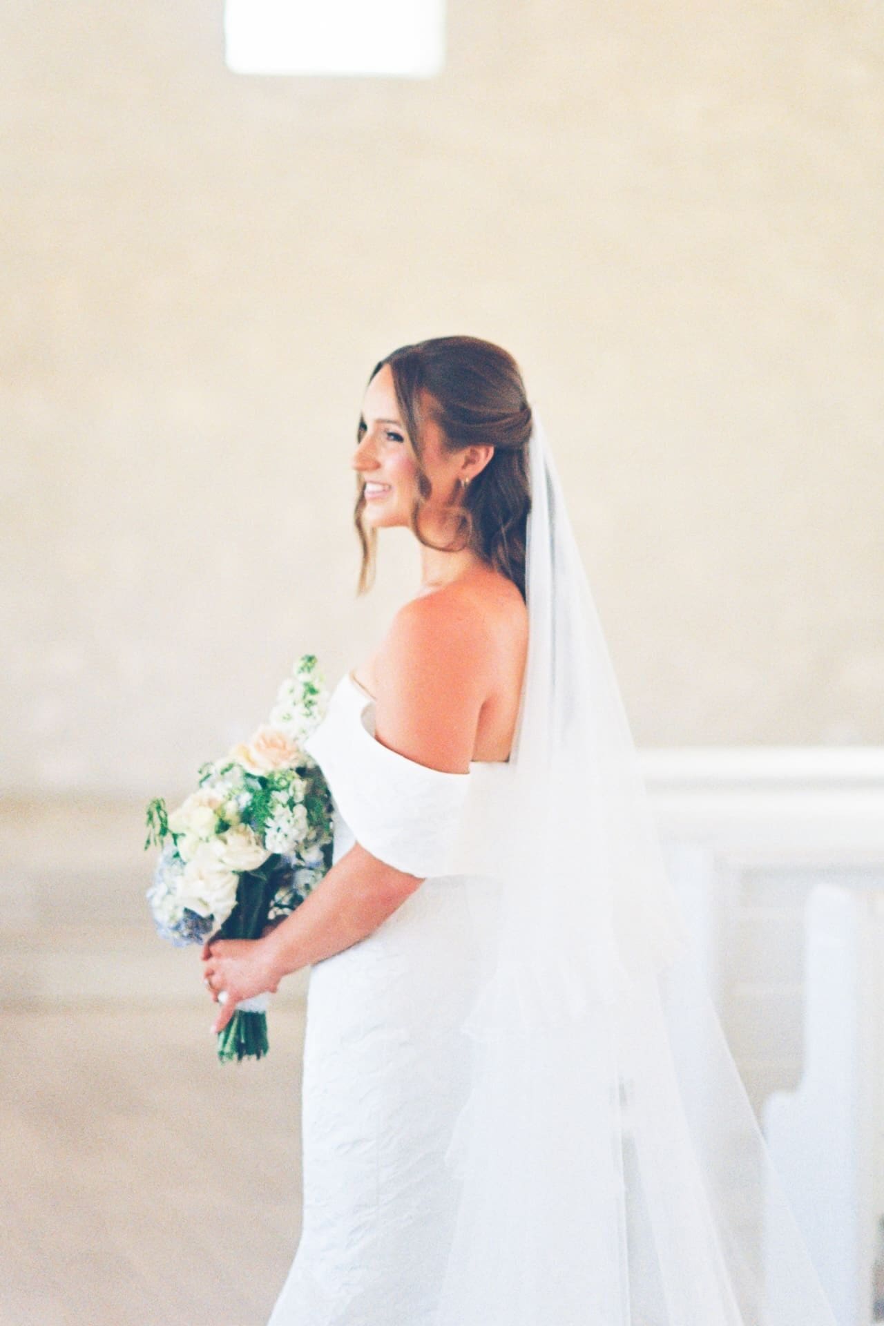 A bride in an off-the-shoulder gown smiles over her shoulder inside a warm stone chapel, her veil trailing behind her as she holds a blue and blush bouquet beside the white pews