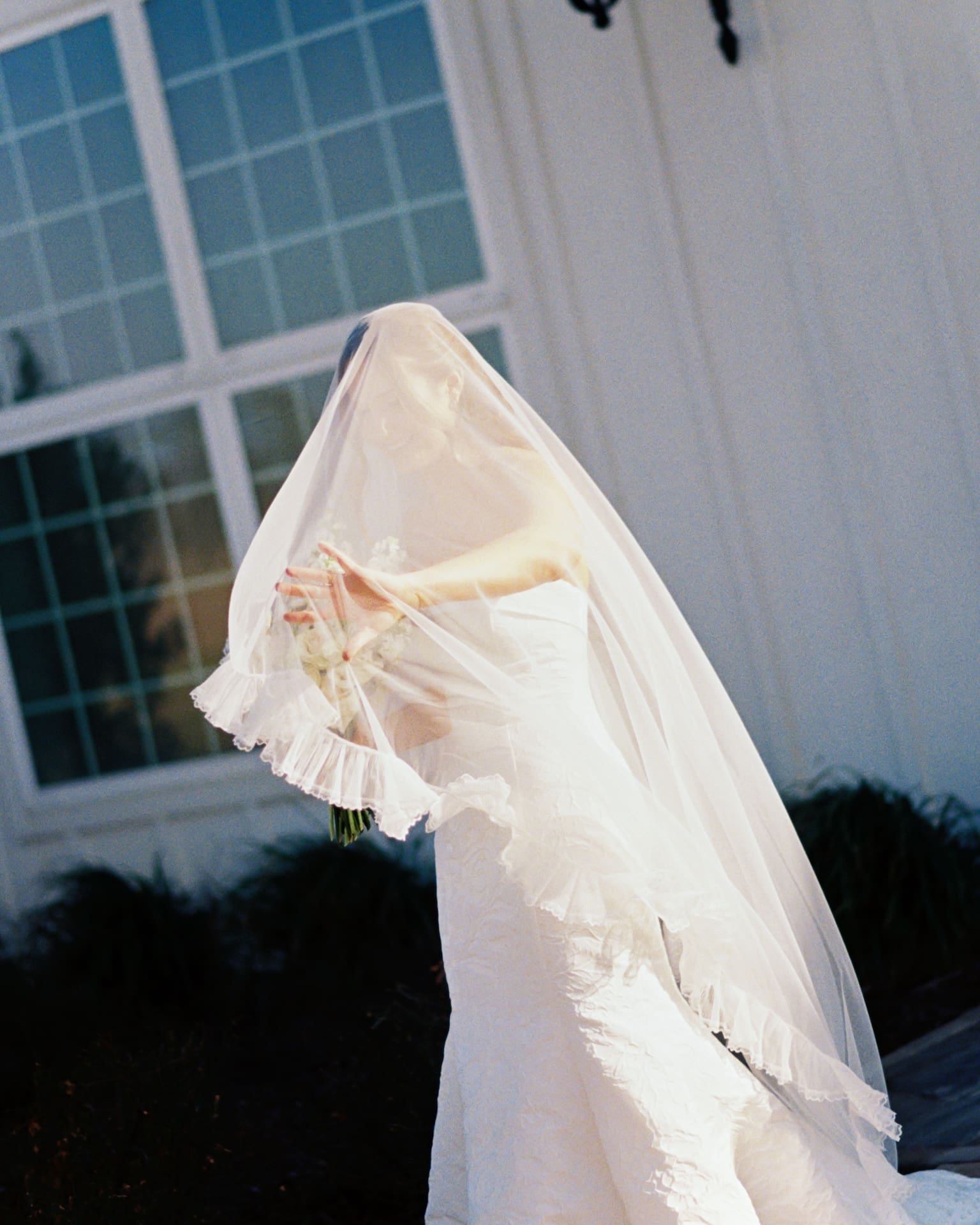Film-style bridal portraits of a bride in a ruffled off-the-shoulder gown standing in soft warm light outside a white chapel, her veil draped over her face as she holds a garden bouquet