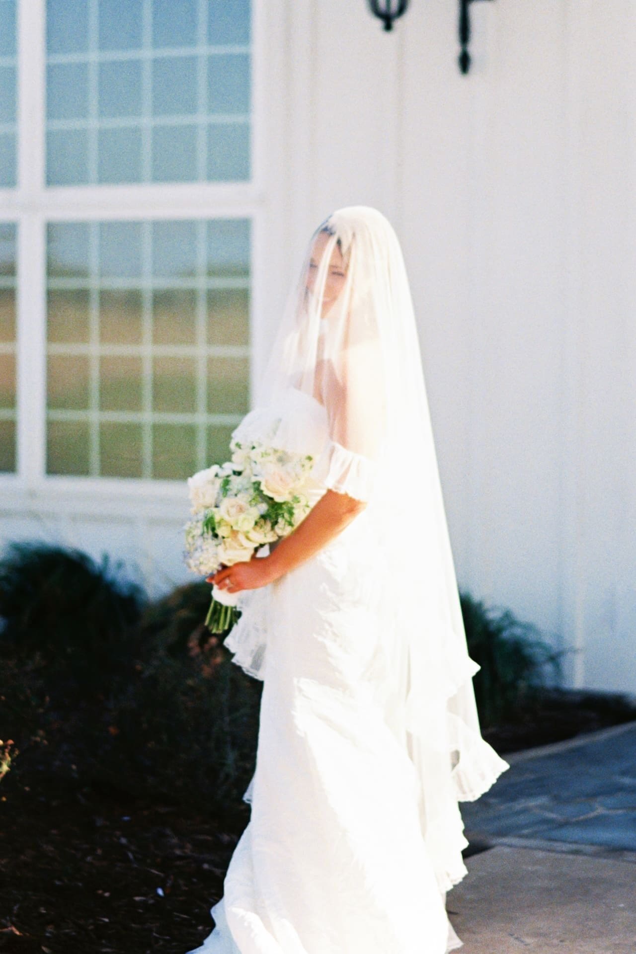 A bride lifts her ruffled veil overhead in golden afternoon light outside a white farmhouse venue, the tulle catching the sun in a luminous, movement-filled moment