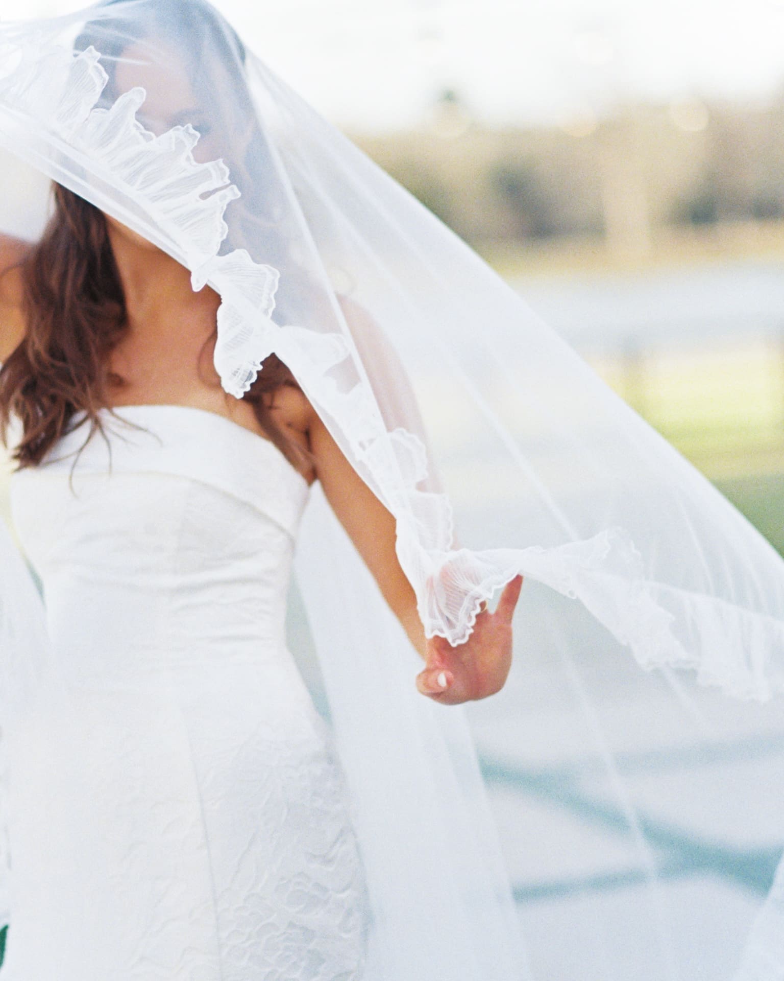 Ethereal bridal portraits captured outdoors as a bride in a strapless lace gown reaches out to catch her ruffled veil as it billows across her face in the breeze