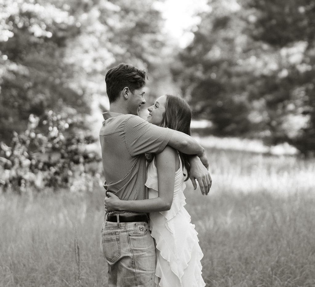 A black and white photo of a couple embracing and smiling at each other in a field, she wears a white ruffle dress and he wears a casual button-down shirt with jeans
