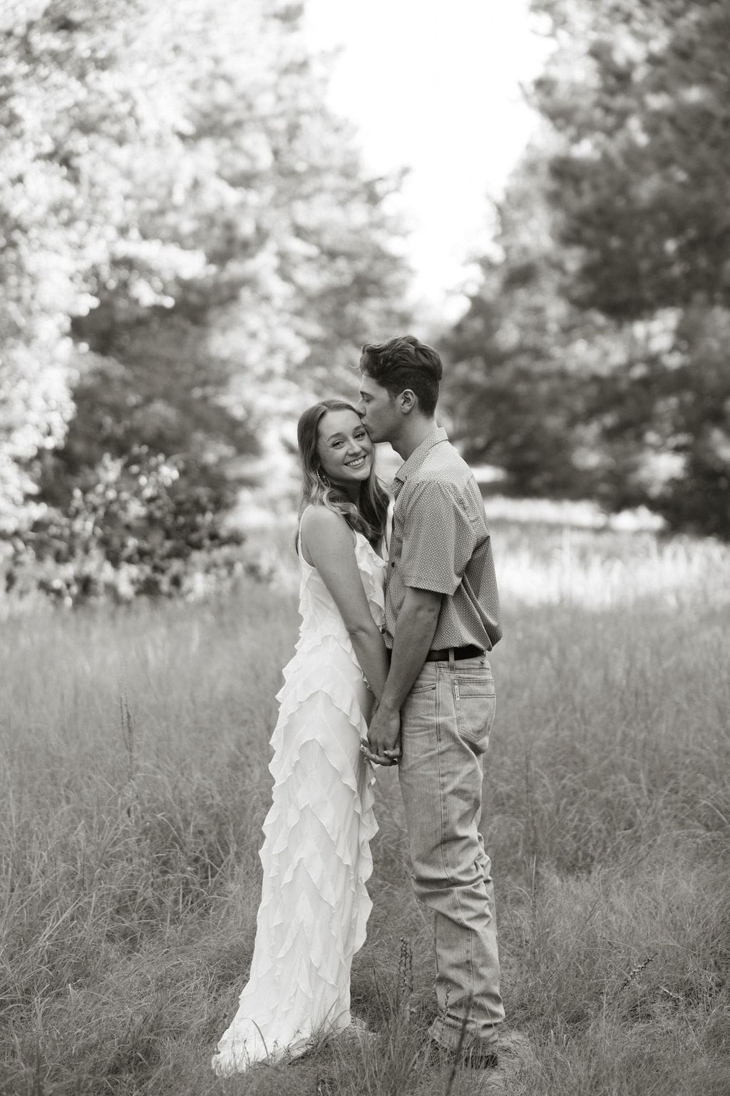 A black and white photo of a couple standing in tall grass, the man kissing her cheek as she smiles over her shoulder, her long white ruffle dress elegant against the soft meadow backdrop