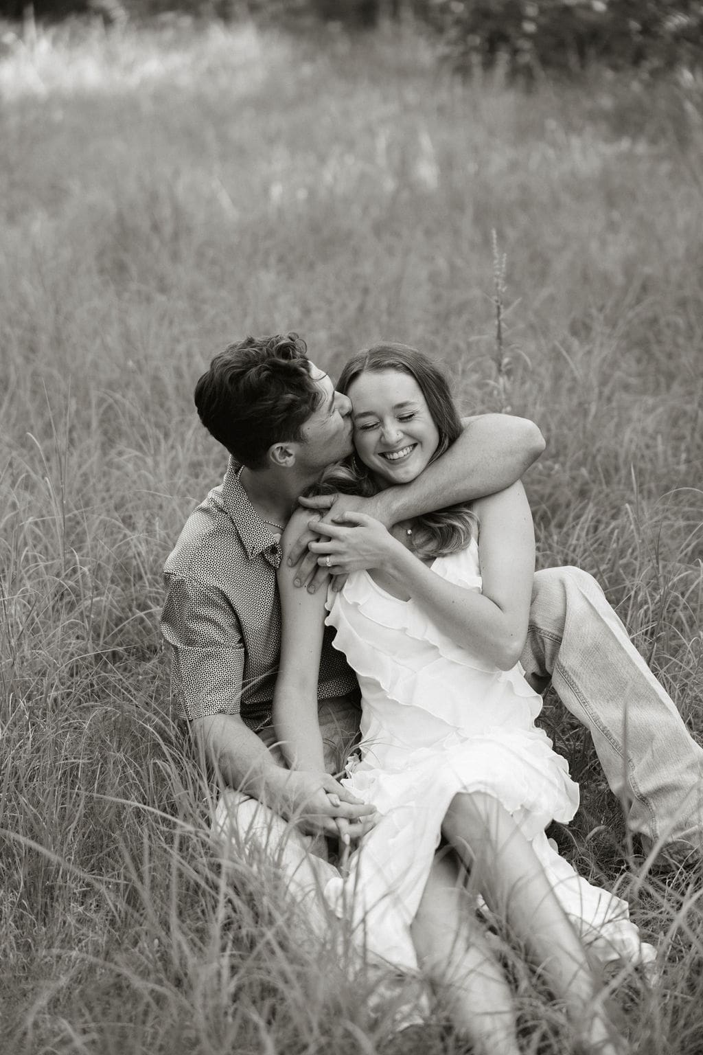A couple sits together in tall grass in black and white, laughing and cuddling as he kisses her temple — she wears a flirty white ruffled dress and he wears a casual button-down shirt