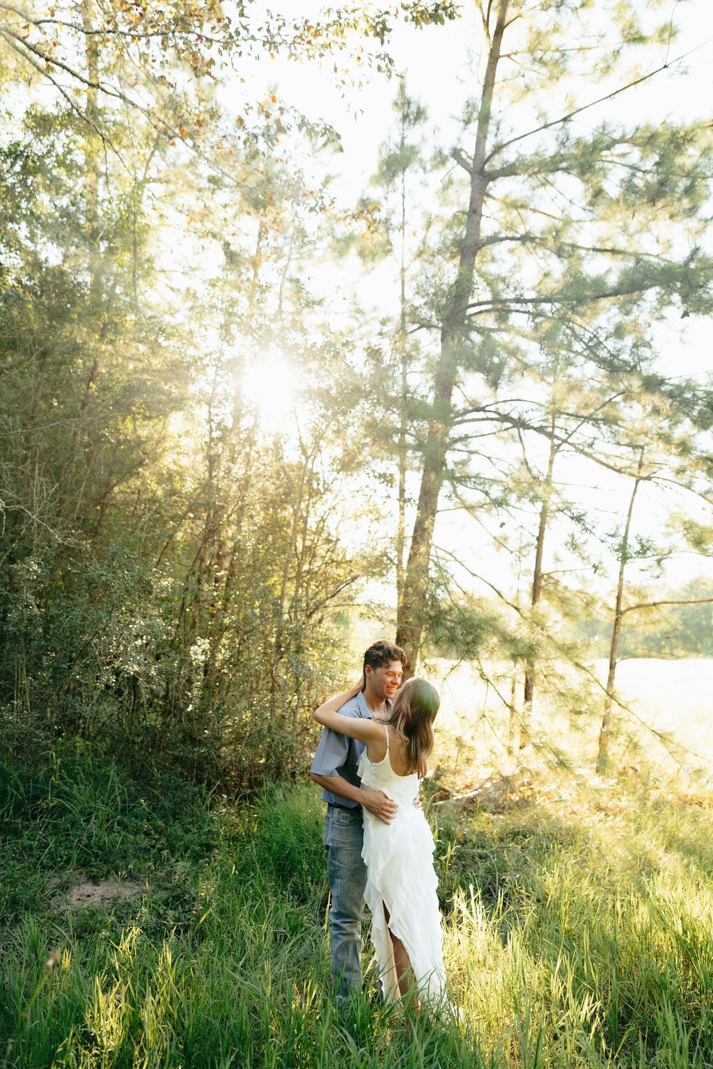 A couple embraces in a sun-filled forest clearing at golden hour, showing what to wear for engagement photos in a natural outdoor setting: a flowing white ruffle dress paired with a blue patterned short-sleeve shirt and jeans