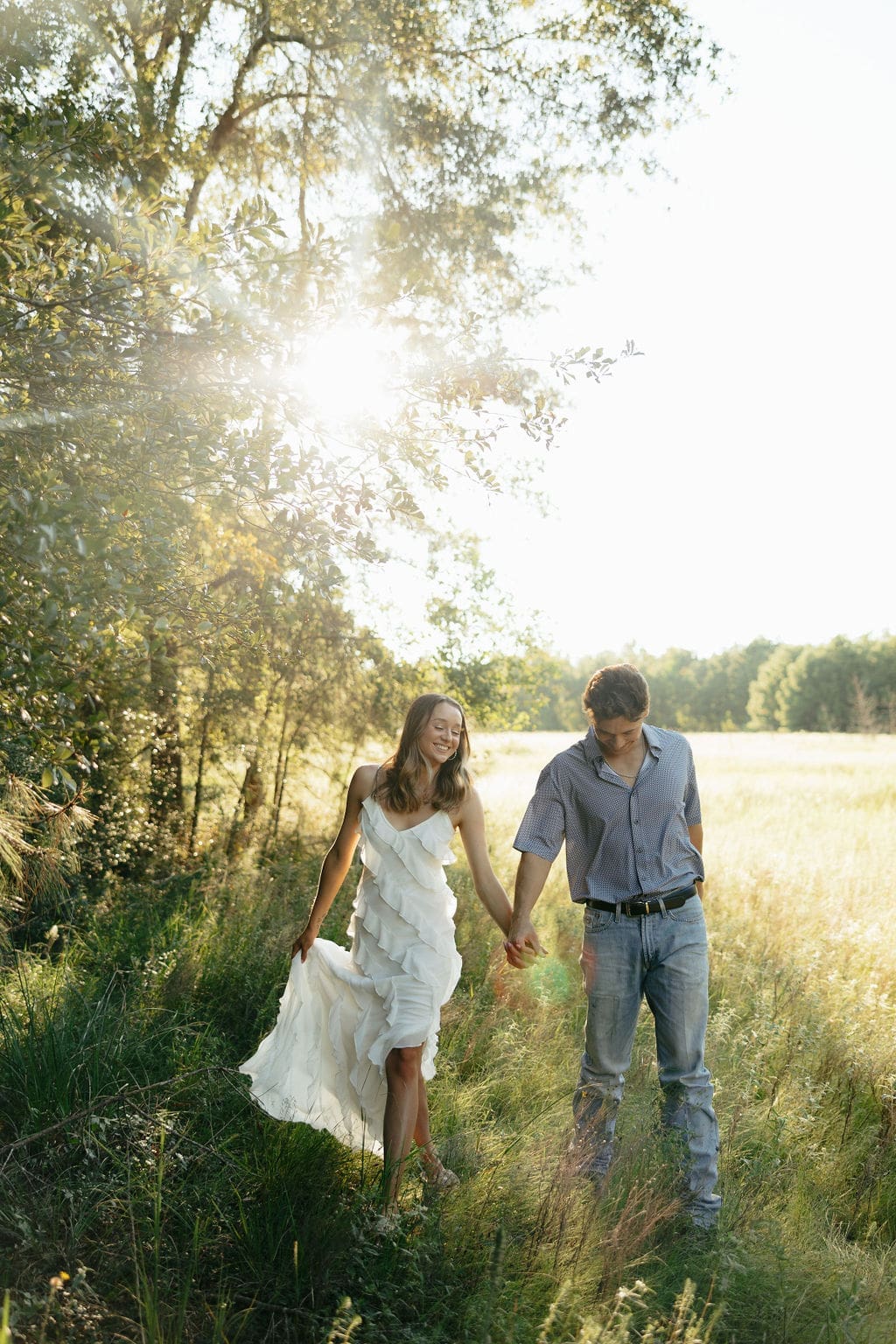 A couple walks hand-in-hand along a sun-soaked field path, showing what to wear for engagement photos in a rustic outdoor setting: a billowing white ruffle dress and a casual blue printed shirt with jeans