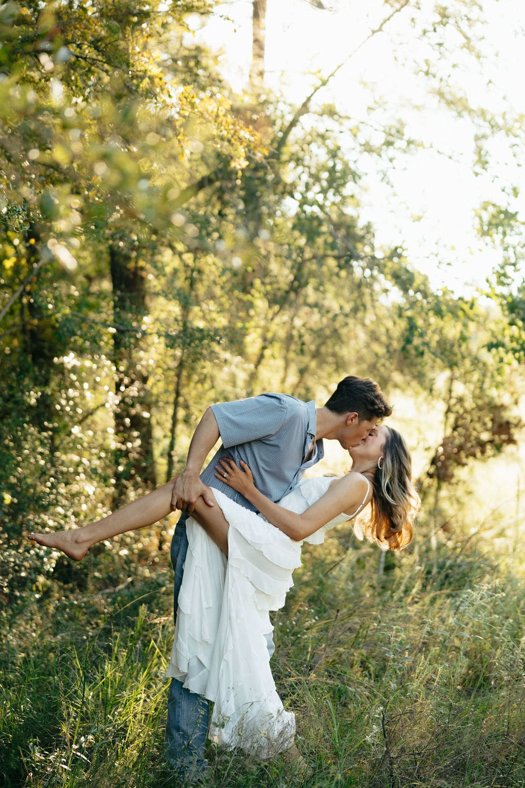 A man lifts and kisses a woman in a sun-drenched forest clearing — what to wear for engagement photos in a wooded setting: a flowing white ruffle dress and a casual patterned button-down create an effortlessly romantic look