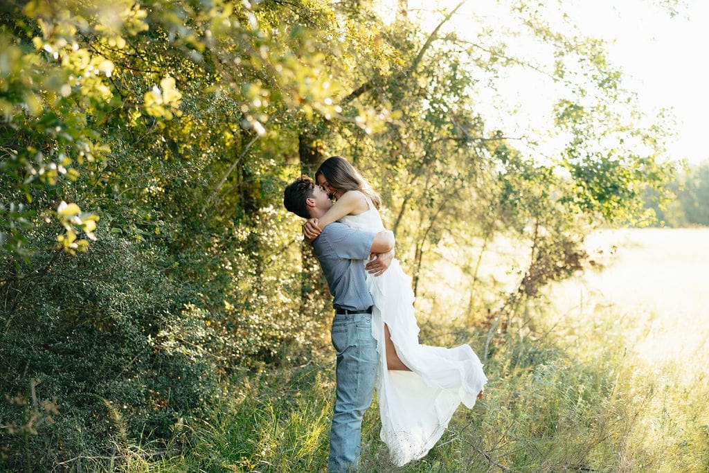 A man lifts and kisses a woman against a glowing wall of trees at golden hour — what to wear for engagement photos in a wooded setting: a flowing white ruffle dress and a blue patterned shirt with jeans create a romantic, natural look