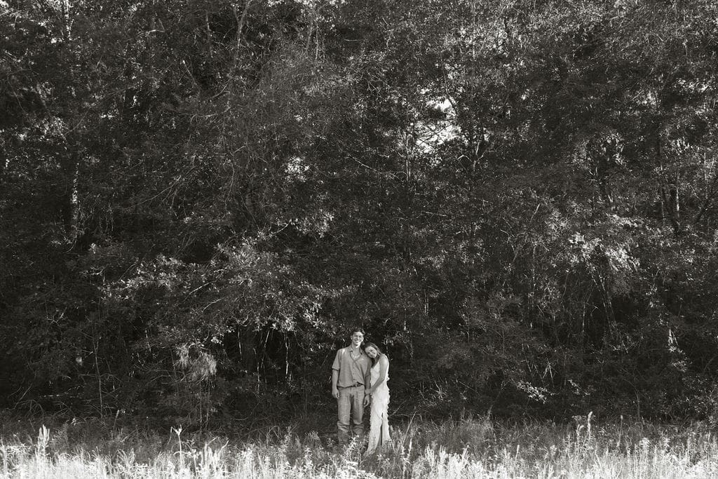 A black and white wide-angle shot of a couple standing at the edge of a lush tree line, leaning into each other in the tall grass and wildflowers
