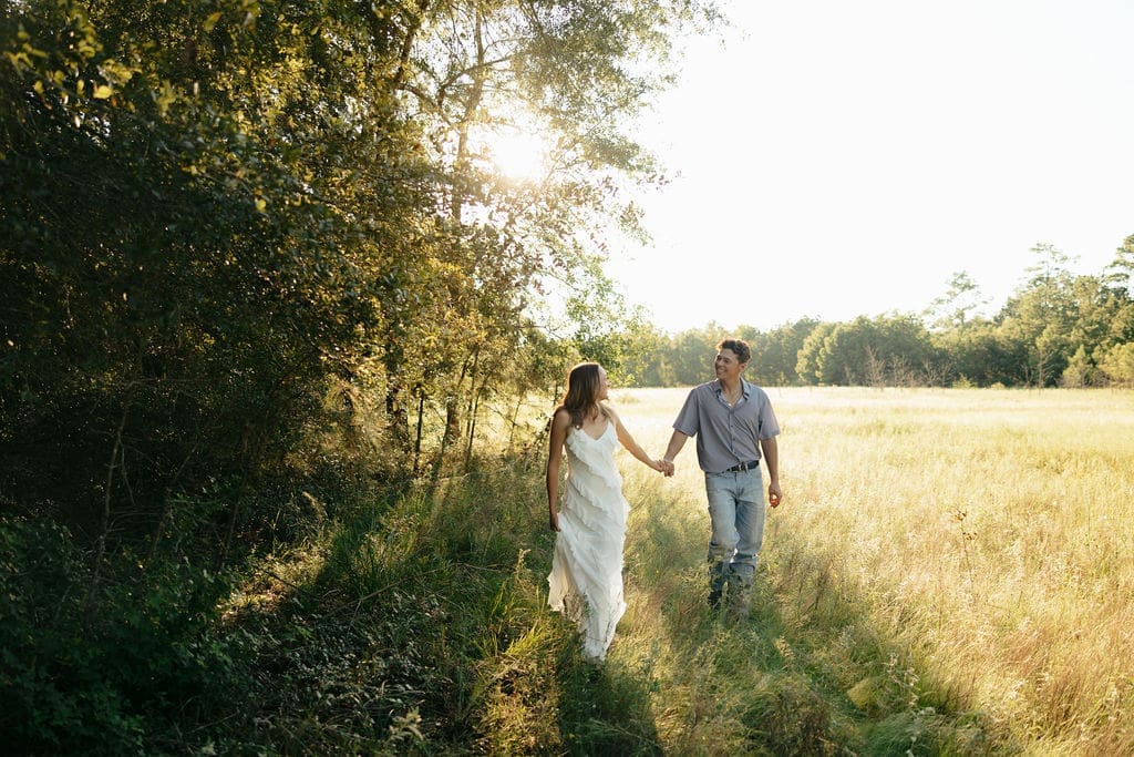 A couple walks together along a sunlit path at the edge of a golden field — what to wear for engagement photos outdoors: a long white ruffle dress paired with a relaxed blue button-down and jeans photographs beautifully in natural evening light