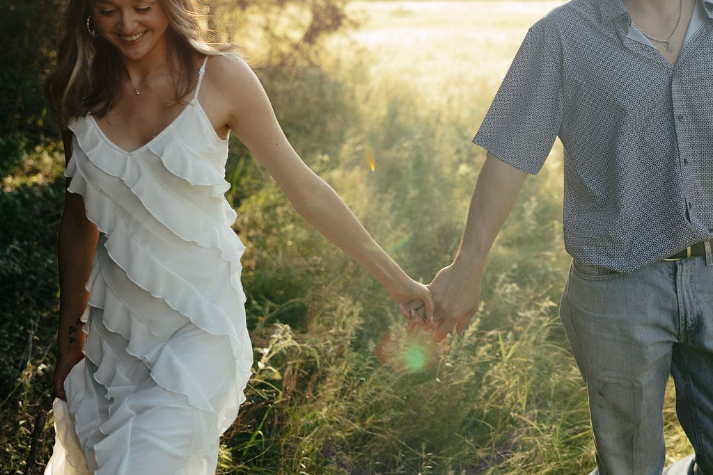 A close-up of a couple holding hands while walking through a sun-drenched field, showing what to wear for engagement photos: a white layered ruffle dress and a casual printed button-down with jeans