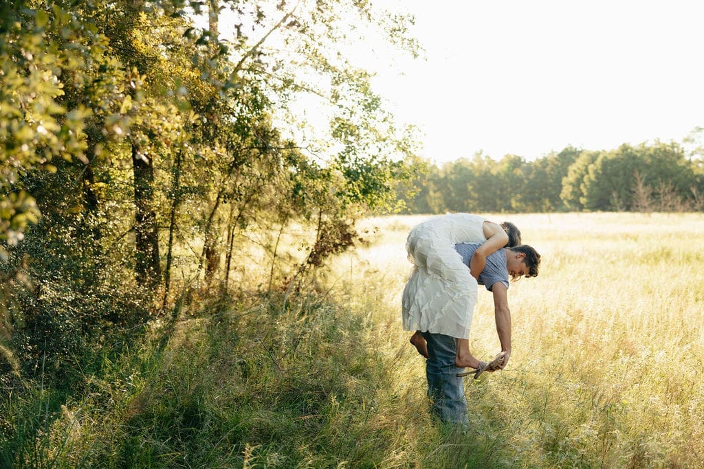 A man playfully carries a woman on his back at the edge of a sun-drenched field, both bent forward and laughing together in the tall grass