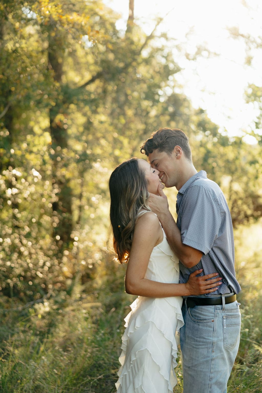 A couple nearly kisses in a glowing golden forest, both smiling with eyes closed, surrounded by soft bokeh light