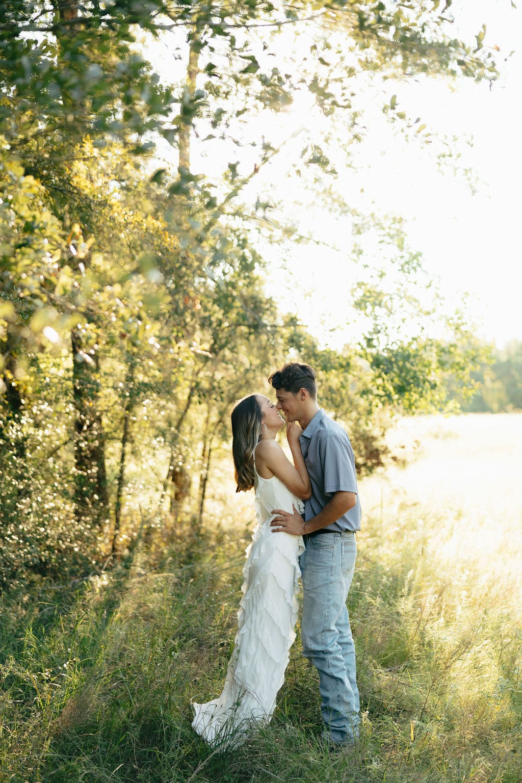 A couple leans in close for a near-kiss in a glowing golden meadow bordered by trees, her white ruffle dress catching the backlight beautifully