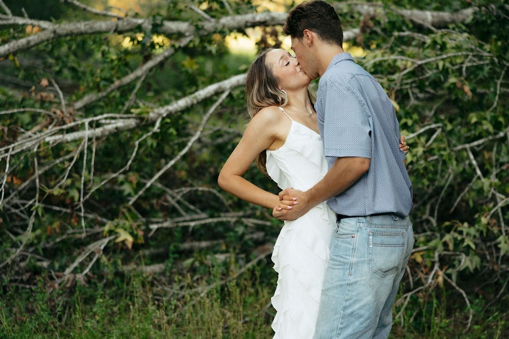 A woman reaches up to kiss a man on the cheek as they stand close together in front of fallen tree branches — what to wear for engagement photos: a white ruffle dress and a relaxed blue short-sleeve shirt keep the look romantic yet laid-back