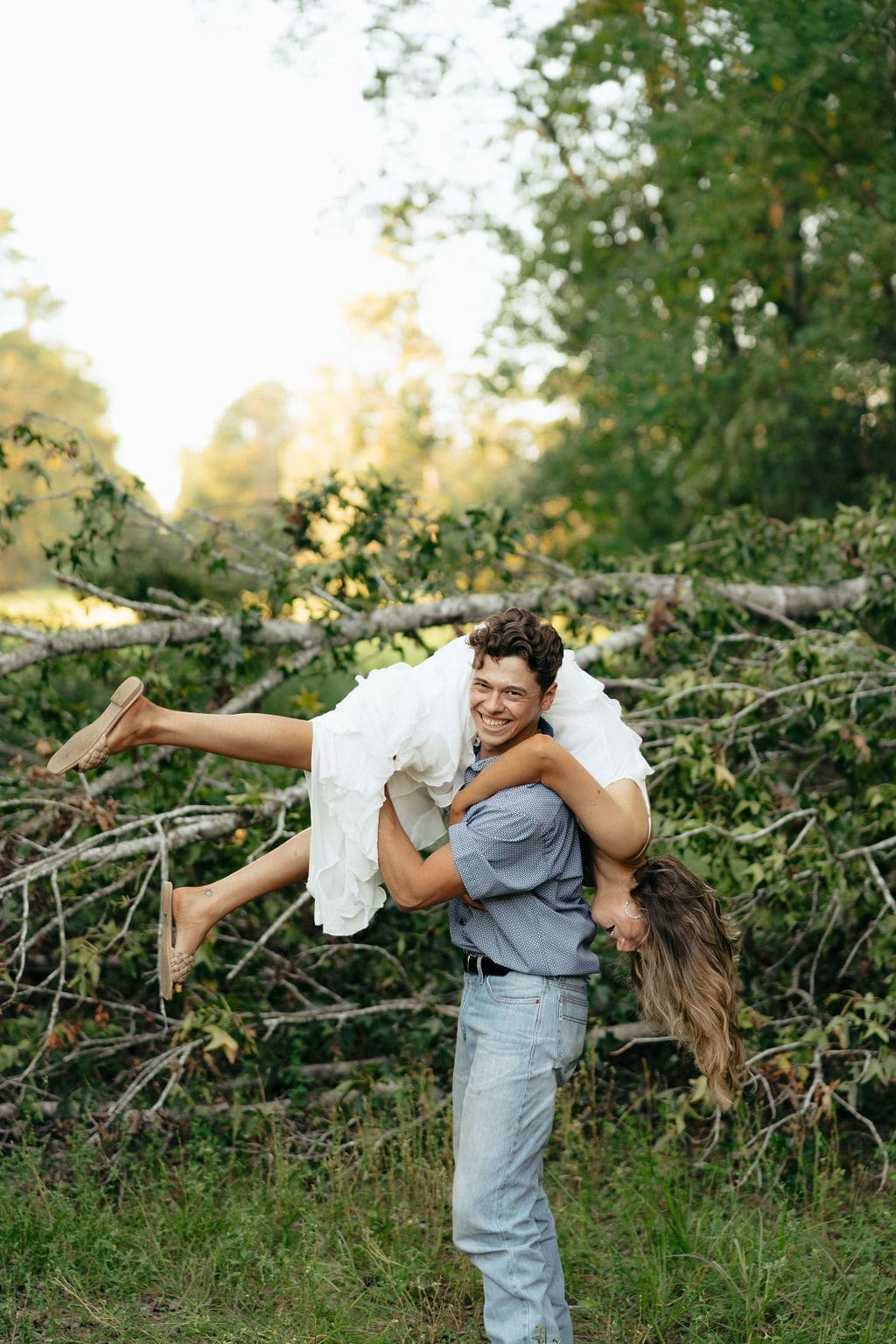 A man laughs as he lifts his partner over his shoulder in front of a pile of fallen trees, both cracking up — she wears a white ruffle dress, he wears a blue patterned shirt and jeans