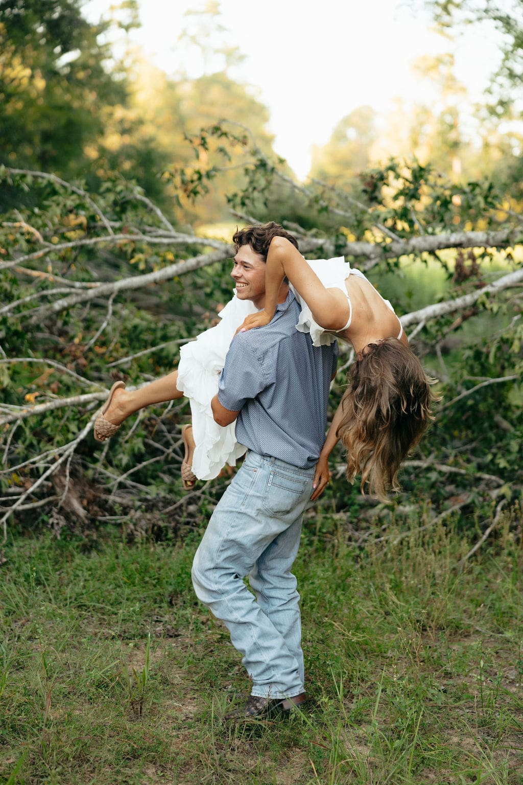 A man laughs and carries his partner upside-down over his shoulder in front of fallen trees, her white ruffle dress flowing — a playful and candid engagement session moment
