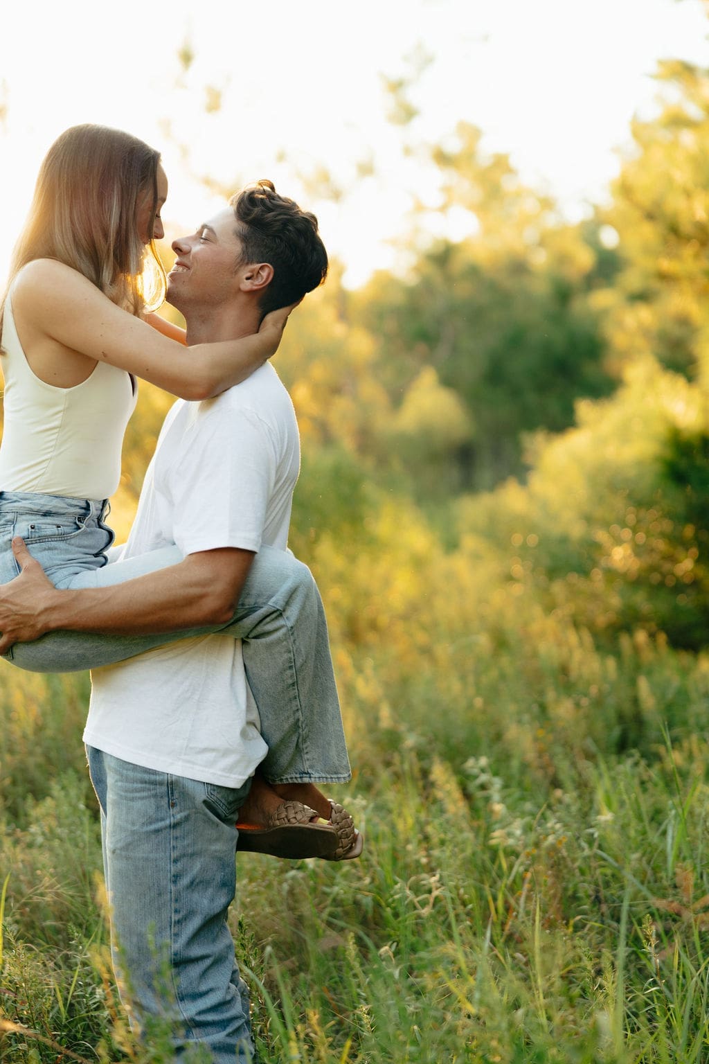 A man lifts a woman in a green wildflower field at sunset, both smiling up at each other — she wears a white tank top and jeans, he wears a white t-shirt and light wash denim