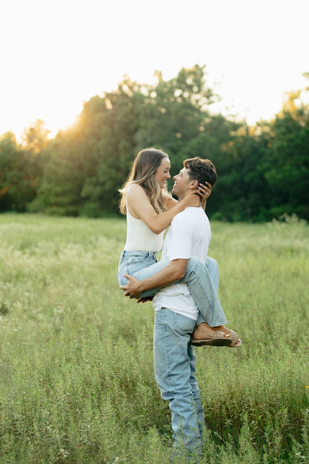 A man lifts a woman in a lush green meadow at golden hour, both gazing at each other and smiling as warm light glows behind the tree line