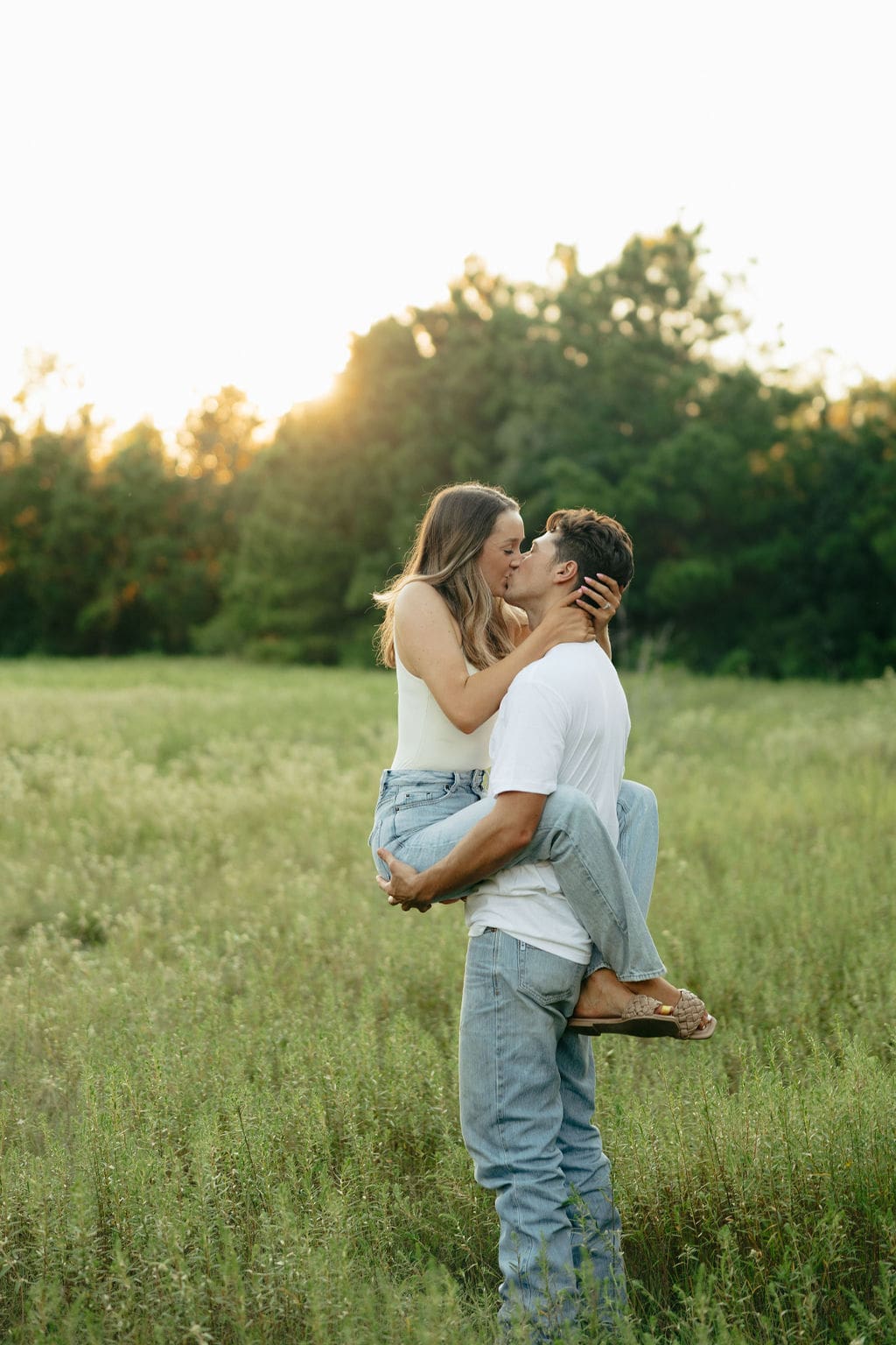A man lifts a woman for a kiss in a lush green meadow at golden hour, her engagement ring visible as she holds his face, both in coordinating white and denim — what to wear for engagement photos: this clean, neutral look photographs beautifully in any natural outdoor setting