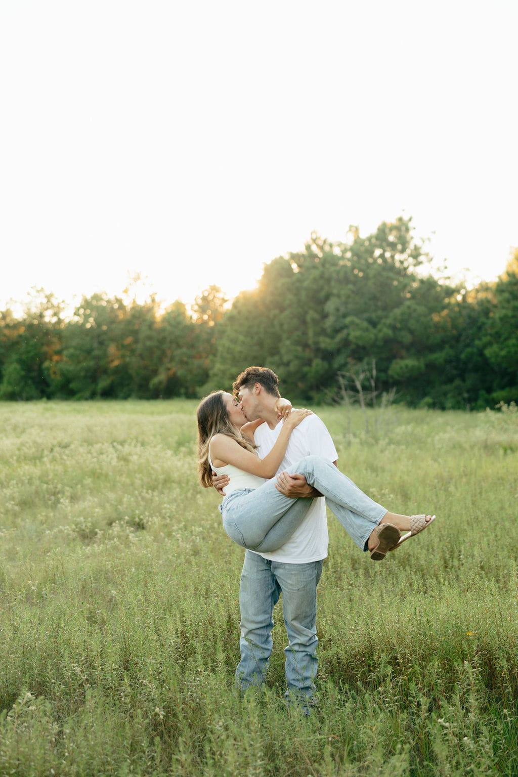 A couple kisses in a sun-drenched meadow at golden hour, the man lifting the woman off her feet — what to wear for engagement photos inspiration: matching light wash jeans and white tops for a timeless, cohesive look