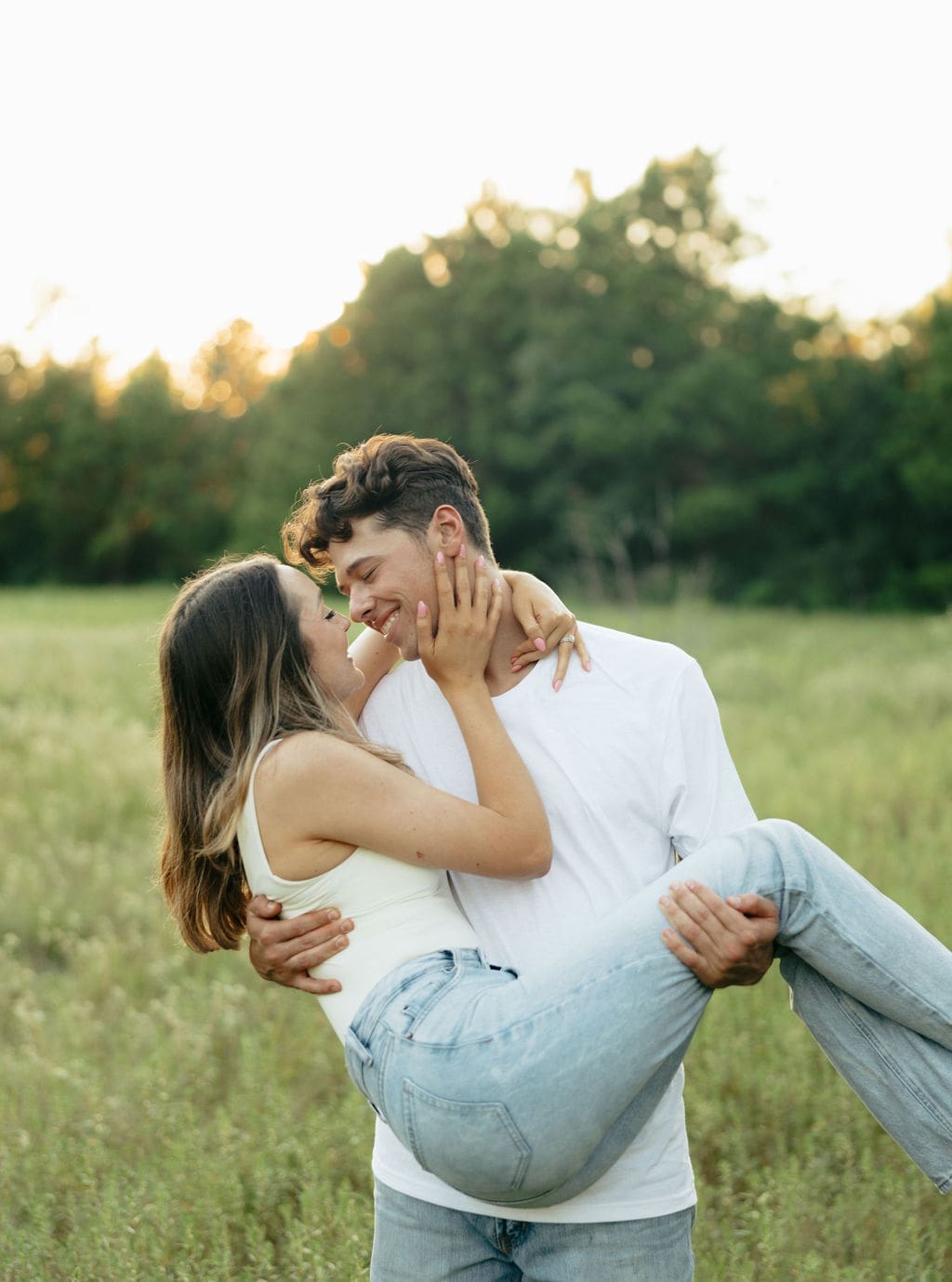 A couple shares a joyful almost-kiss as he carries her in a green meadow at dusk — what to wear for engagement photos: coordinating white and neutral tones keep the focus on the connection between them