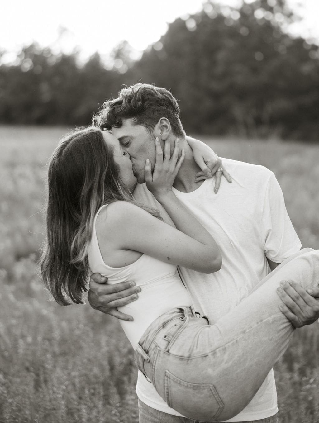 A close-up black and white photo of a couple sharing a passionate kiss in a field, the woman's engagement ring visible as she holds his face