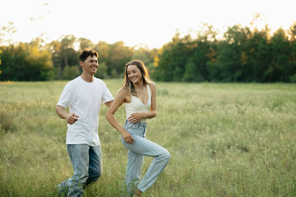 A couple laughs and walks together through a lush green meadow at golden hour, both in matching white tops and light wash jeans