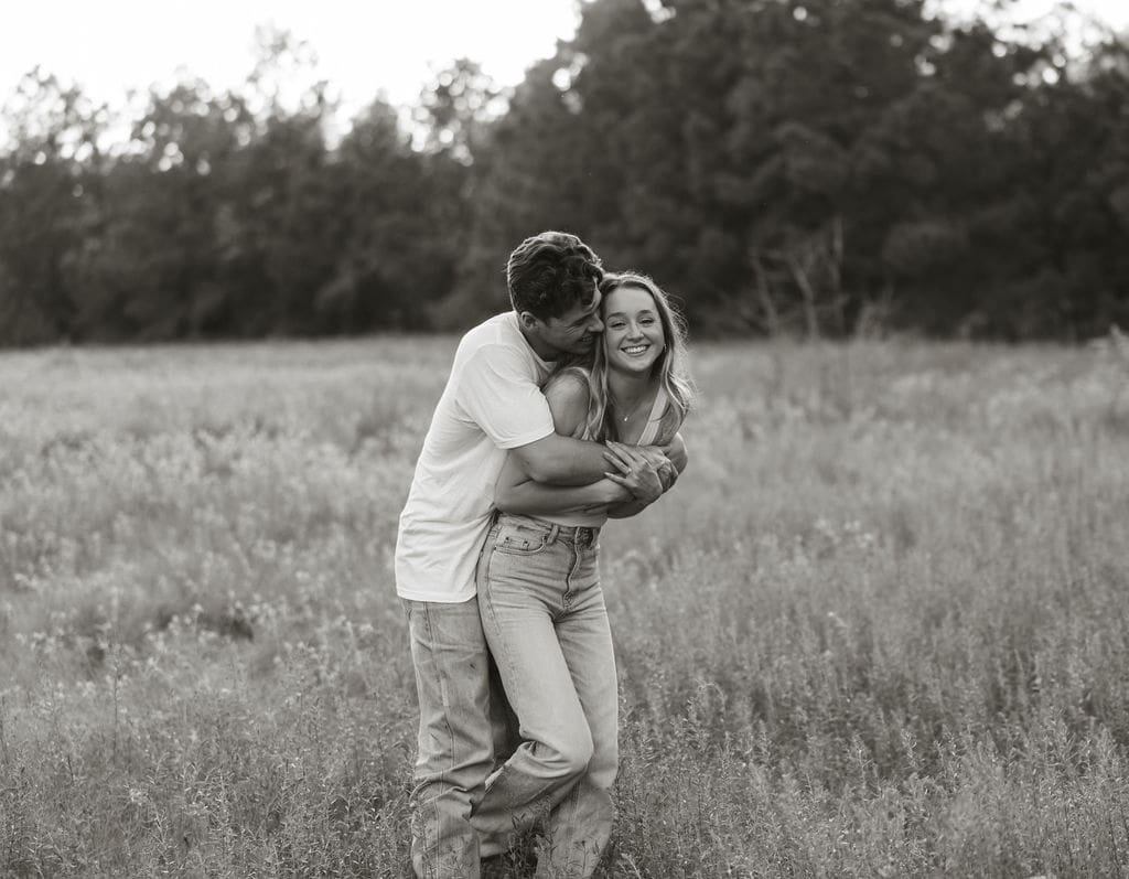 A black and white photo of a man hugging a woman from behind in a wildflower field, both laughing as the treeline blurs softly in the background — what to wear for engagement photos: simple white and neutral tones let the emotion and connection take center stage