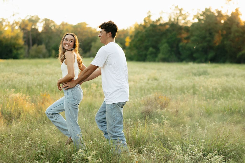 A couple laughs and walks together through a lush green meadow at golden hour, both in matching white tops and light wash jeans