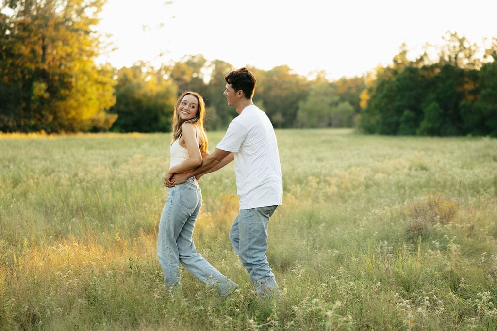 A couple dances together in a golden meadow at sunset, the woman glancing back over her shoulder with a bright smile — what to wear for engagement photos: coordinating white tops and light wash jeans are a classic, effortless choice for an outdoor session