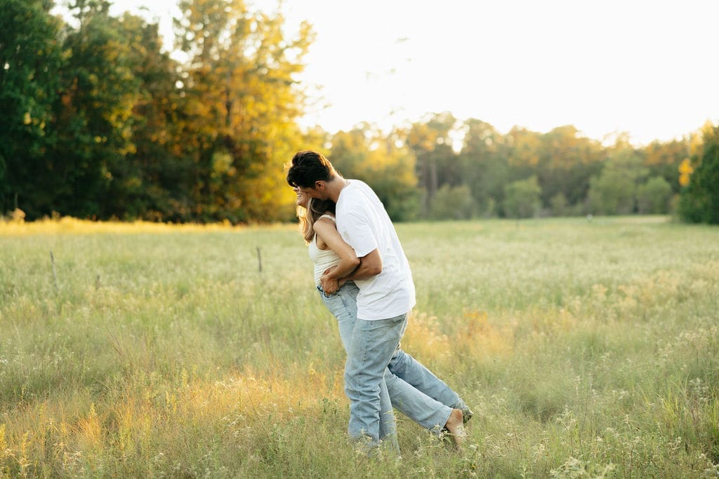 A couple playfully embraces in a golden meadow at sunset, the man wrapping his arms around her from behind and nuzzling her neck — what to wear for engagement photos: simple white and neutral tones photograph beautifully in natural light