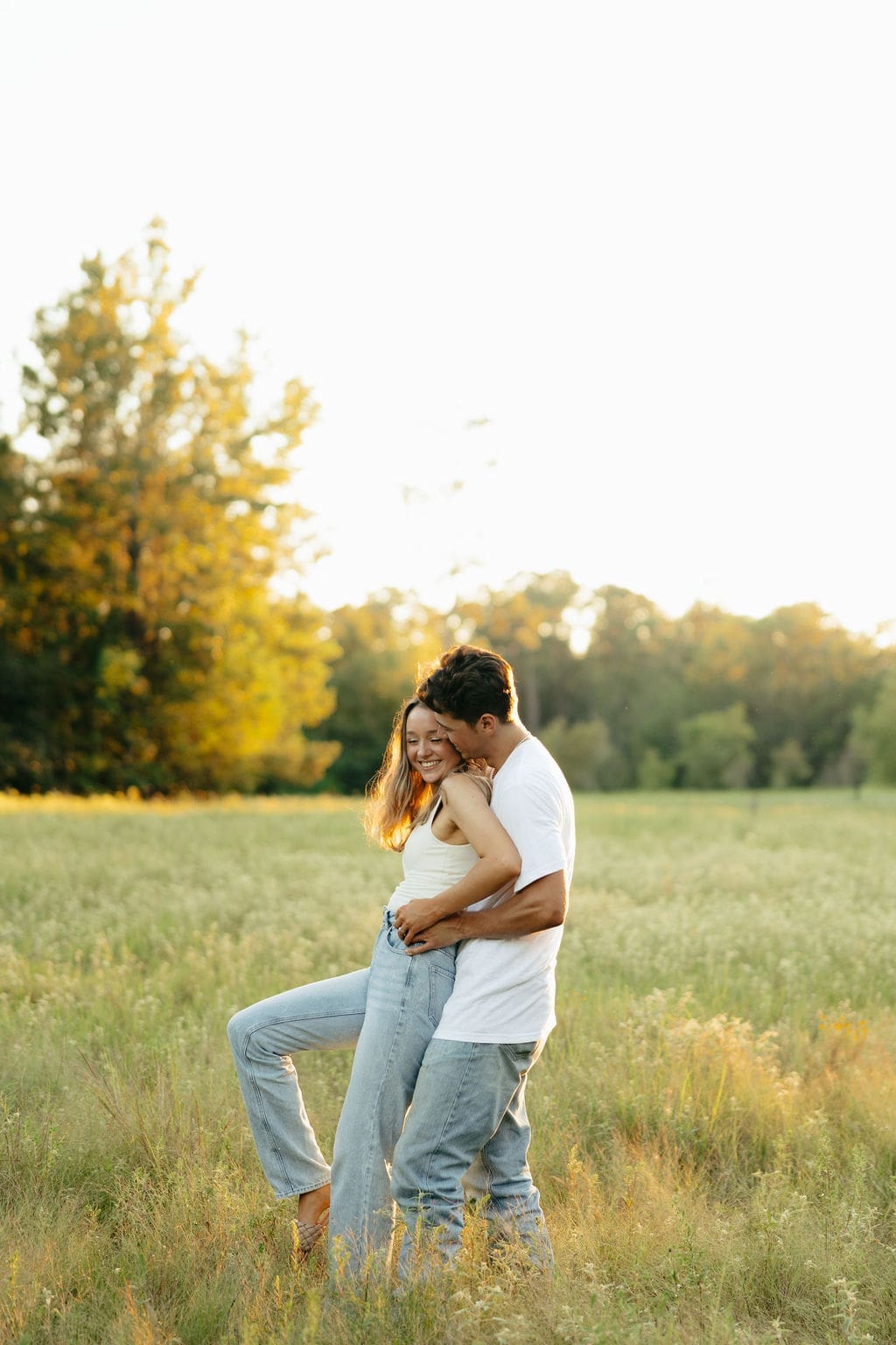 A couple laughs together in an open field at sunset, the man kissing her neck from behind as she smiles with her leg kicked up