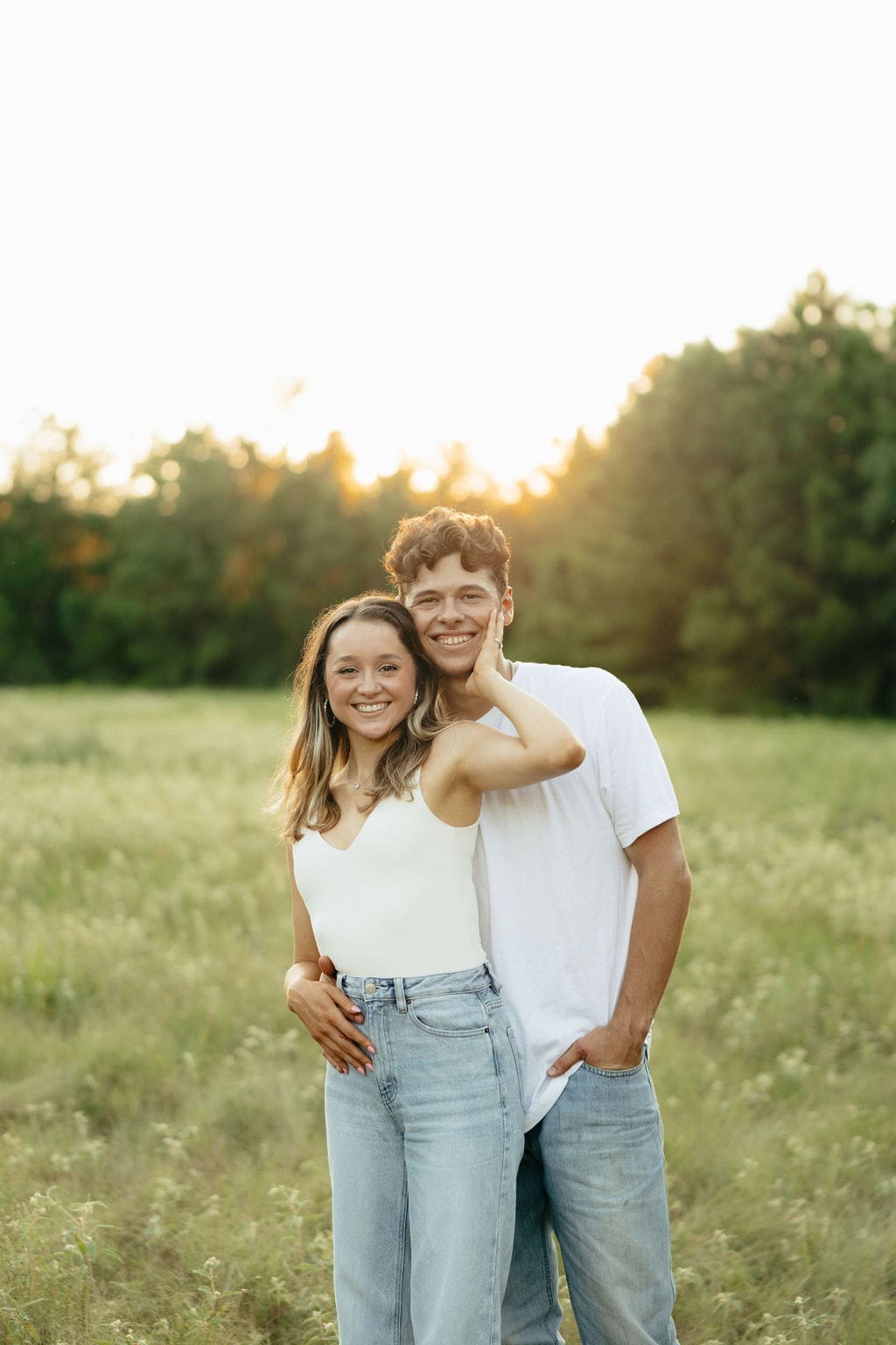 A couple stands close together in a green meadow at golden hour, both smiling at the camera in coordinating white tops and light wash jeans as warm light glows behind them