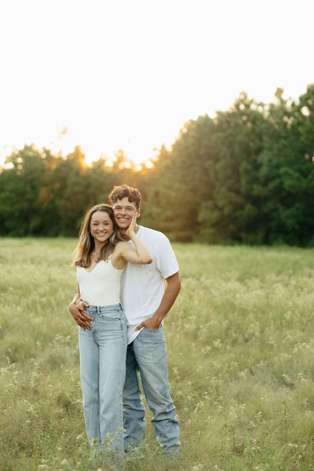 A couple smiles warmly at the camera in a green meadow at golden hour, both wearing coordinating white tops and light wash jeans with the sun glowing through the trees behind them