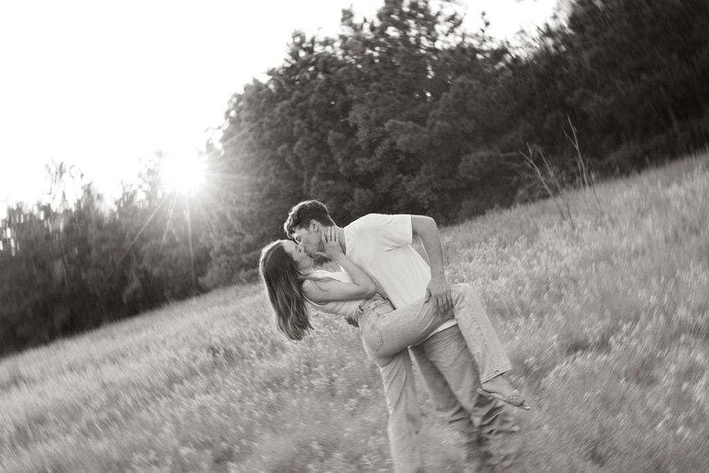 A dramatic black and white photo of a couple kissing mid-lift in a field with a sunburst bursting through the tree line behind them