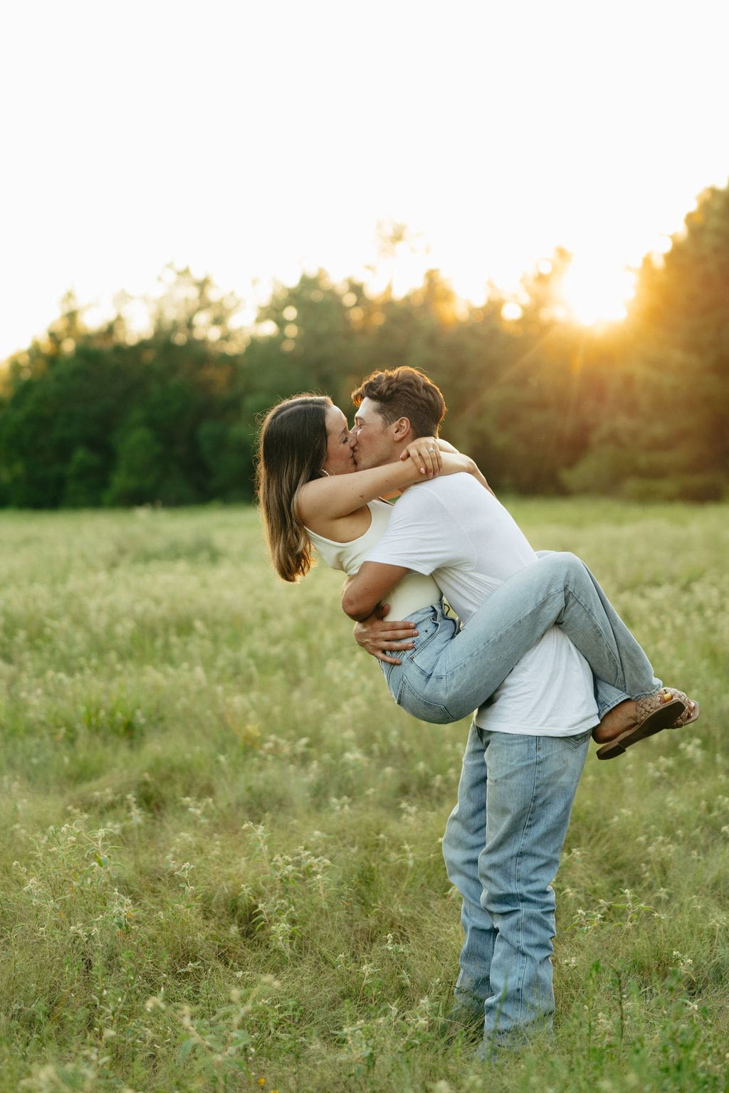 A man lifts and kisses a woman in a wildflower field at sunset, both in coordinating white tops and light wash jeans with a warm golden glow behind the trees — what to wear for engagement photos: neutral tones and denim are a go-to combination for outdoor sessions