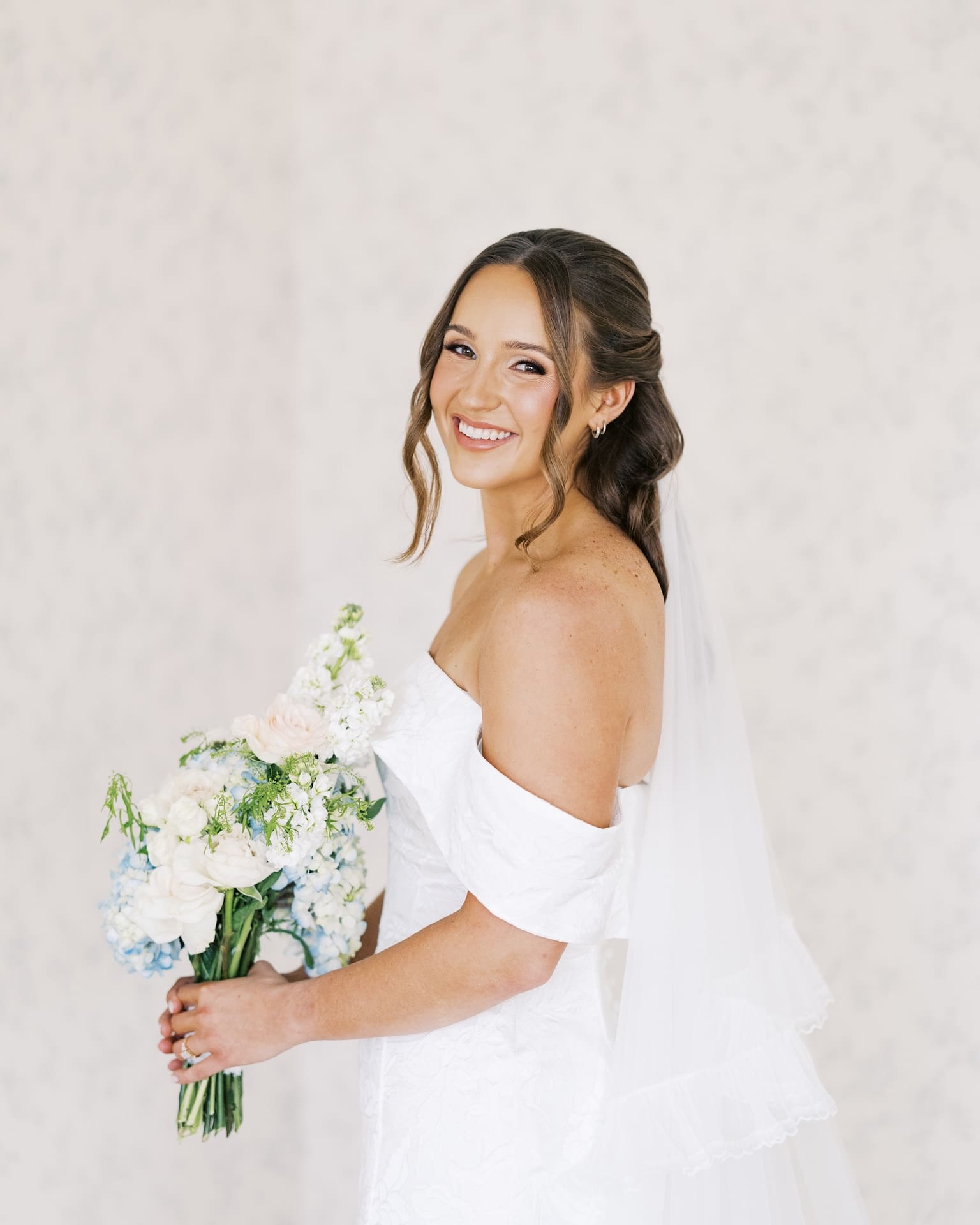 A bride in an off-the-shoulder gown beams over her shoulder while holding a blue hydrangea and white rose bouquet against a soft neutral backdrop