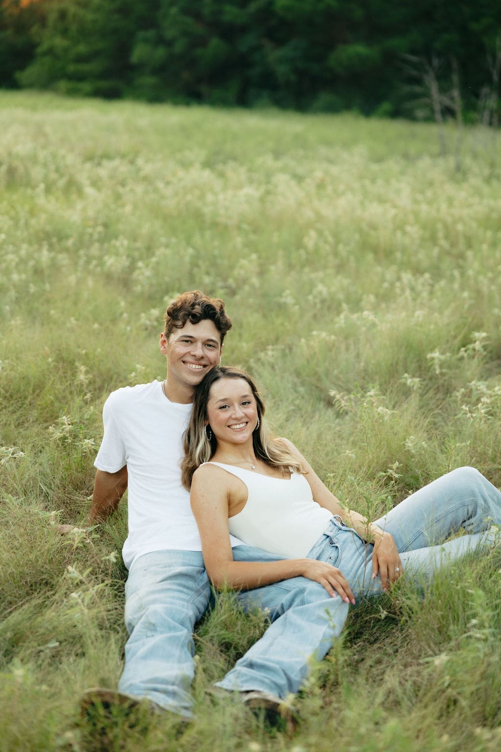 A couple sits together in a wildflower field smiling at the camera, both in white tops and light wash jeans, relaxed and happy in the tall grass