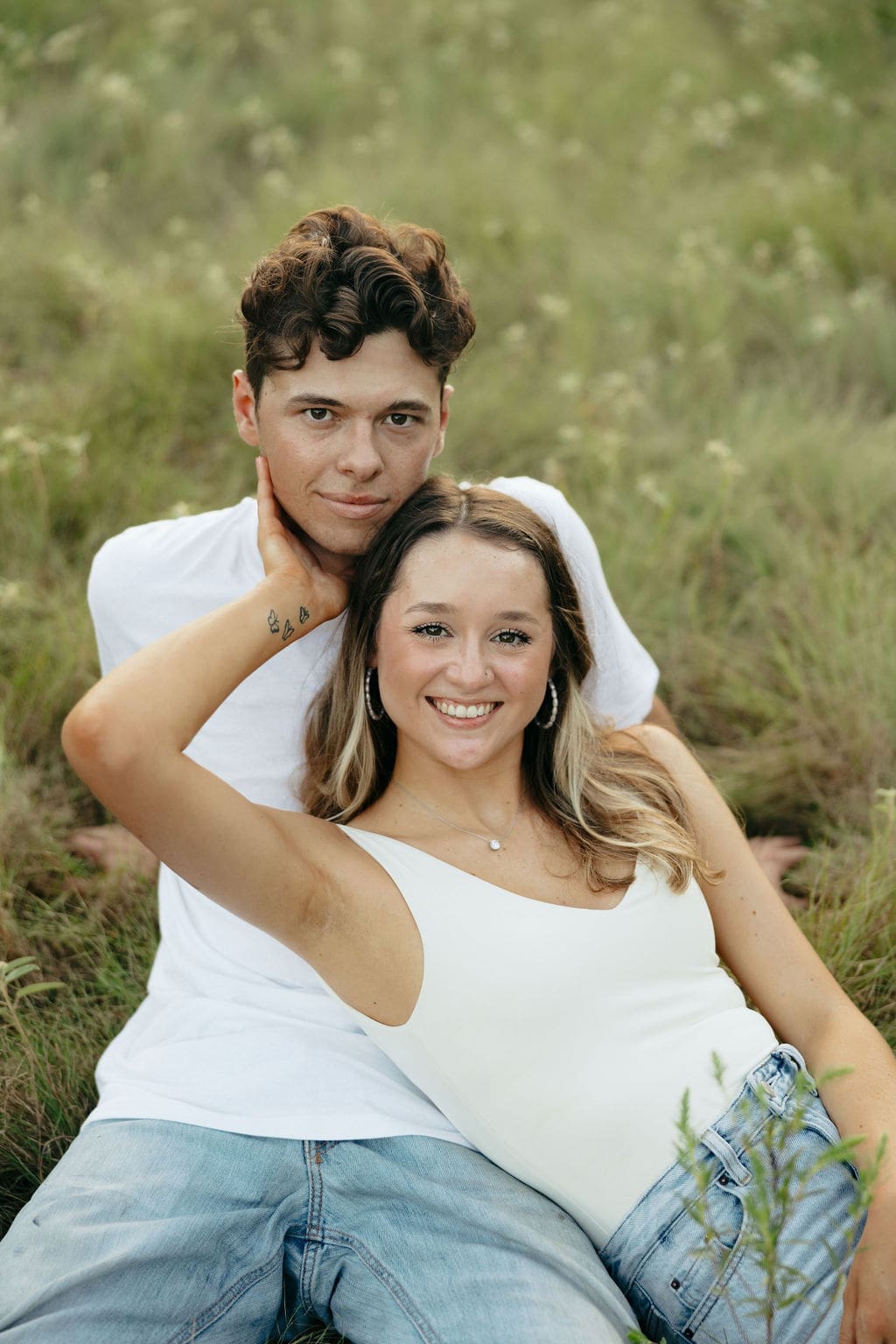 A couple sits together in tall grass smiling at the camera — what to wear for engagement photos: matching white tops and light wash jeans create a clean, coordinated look perfect for an outdoor session