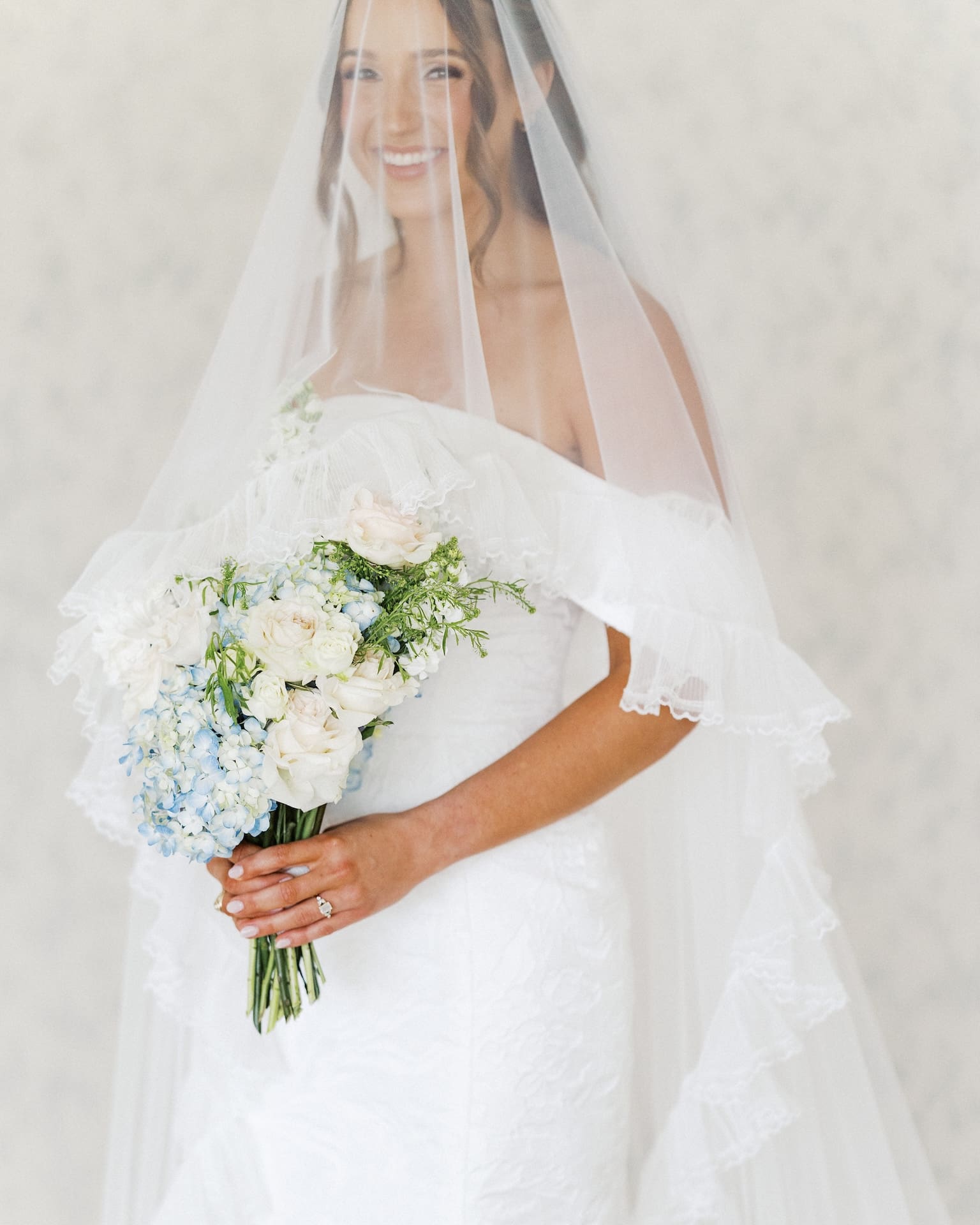 Close-up bridal portraits of a bride holding a blue hydrangea and white rose bouquet, her ruffled veil framing her face as she smiles in front of a neutral textured wall