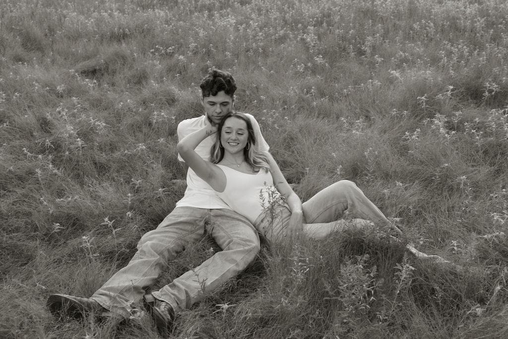 A black and white photo of a couple relaxing together in tall grass, she leans back against him and smiles softly as he wraps his arm around her