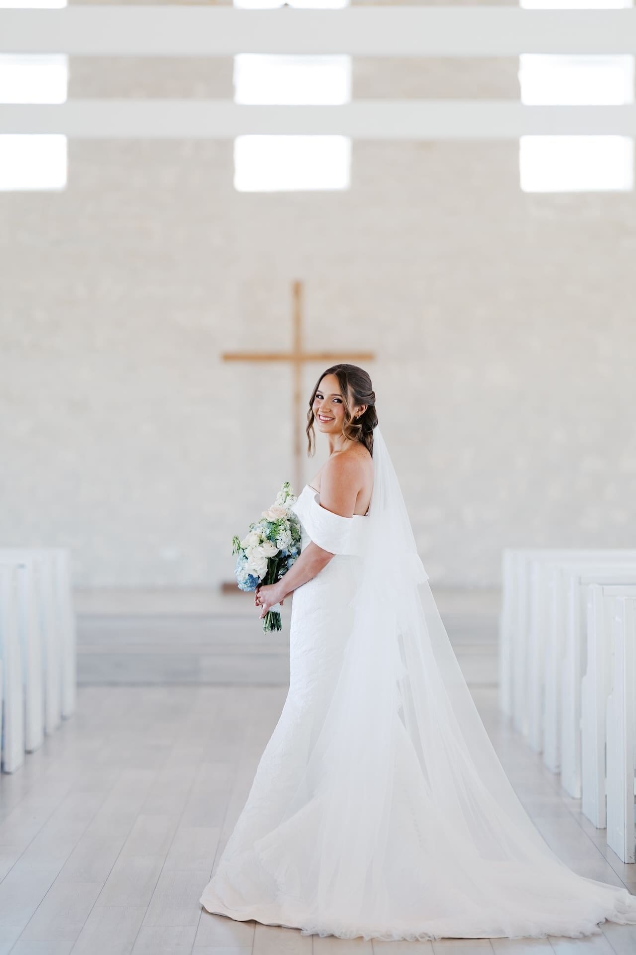 A bride in an off-the-shoulder gown and cathedral veil glances back over her shoulder with a radiant smile, standing in the aisle of a bright modern chapel with a wooden cross behind her