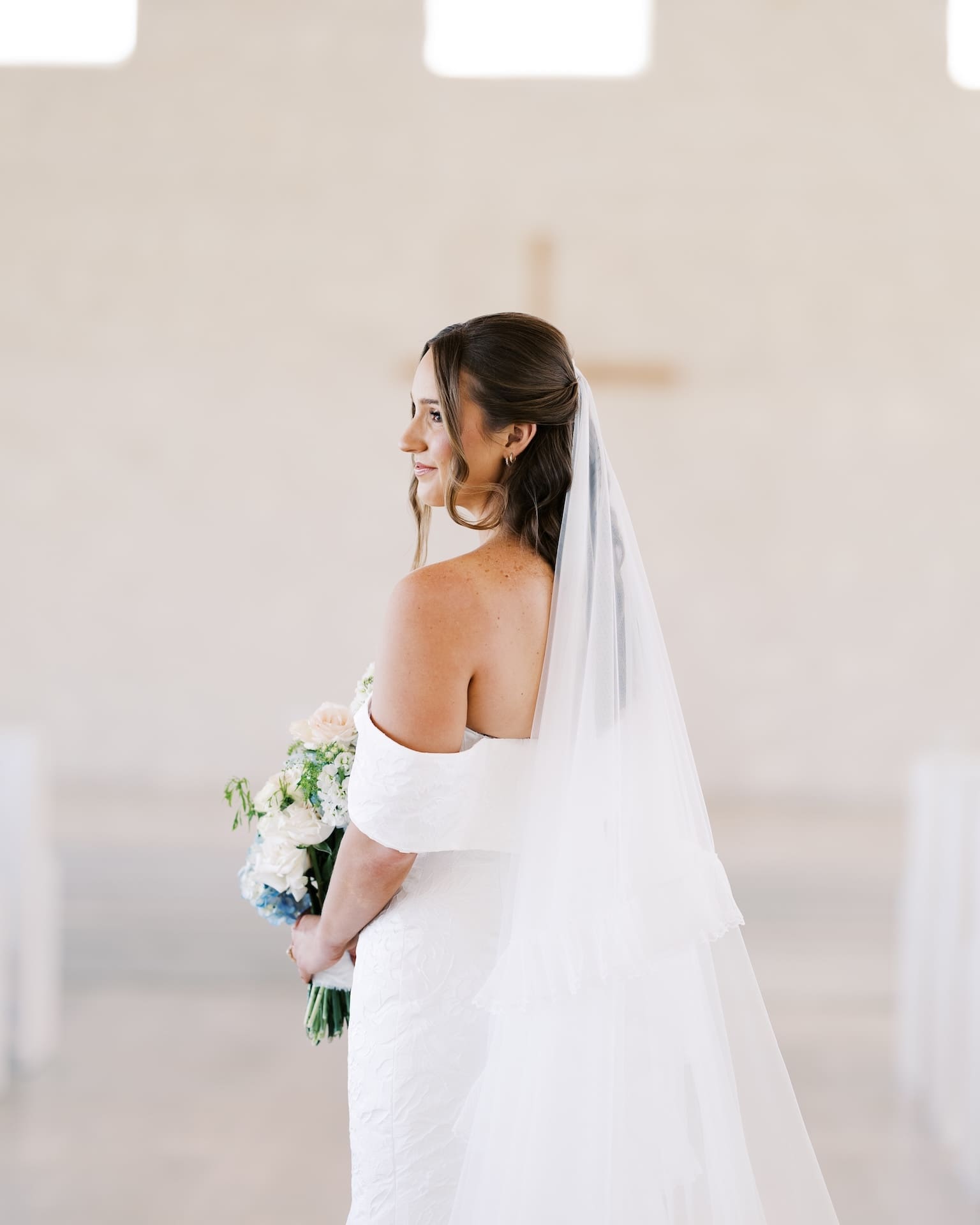 Serene bridal portraits of a bride in an off-the-shoulder gown standing with her back to the camera inside a white chapel, a wooden cross softly blurred behind her