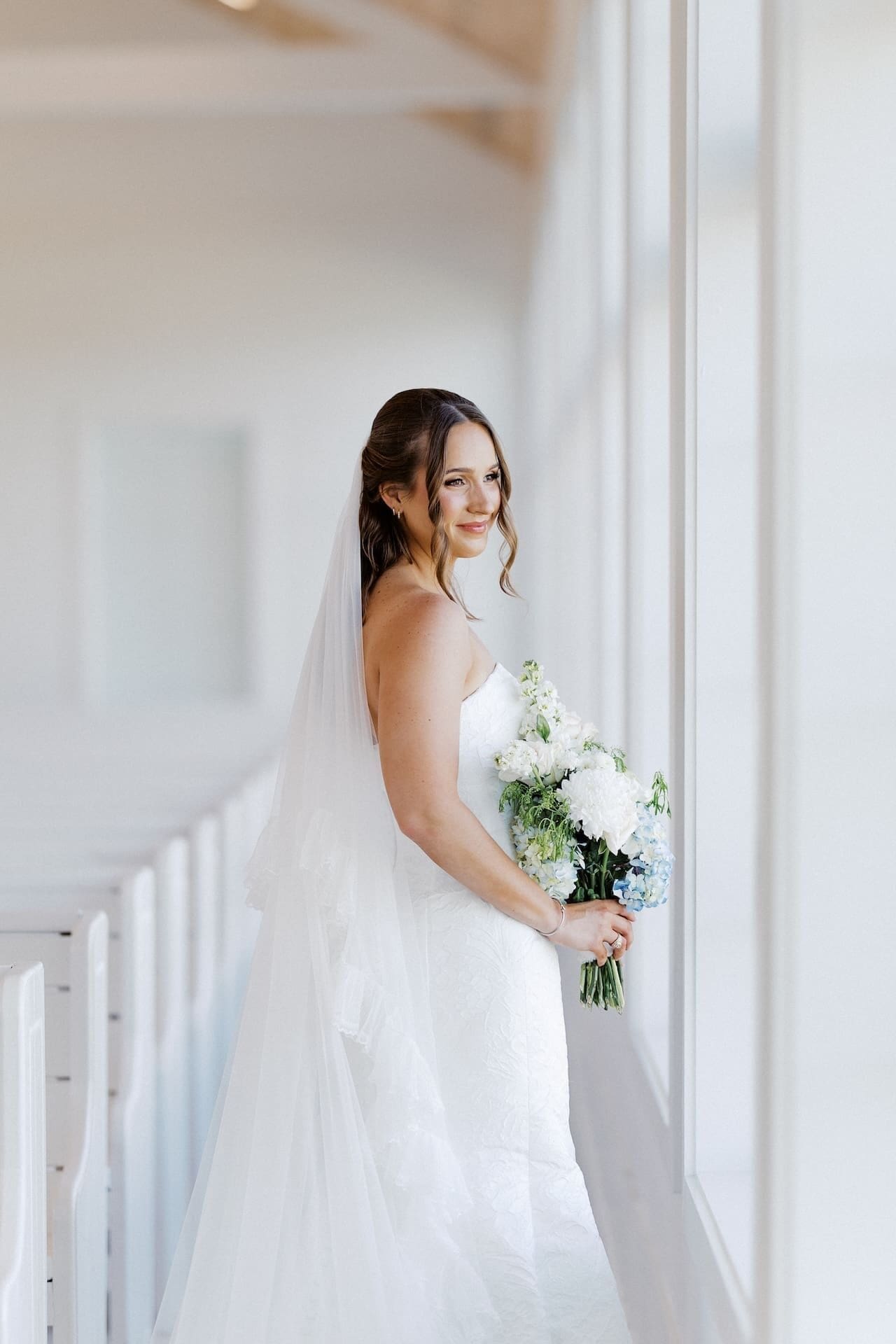 A bride stands by a tall window in a bright hallway, smiling back over her shoulder while holding a blue and white bouquet