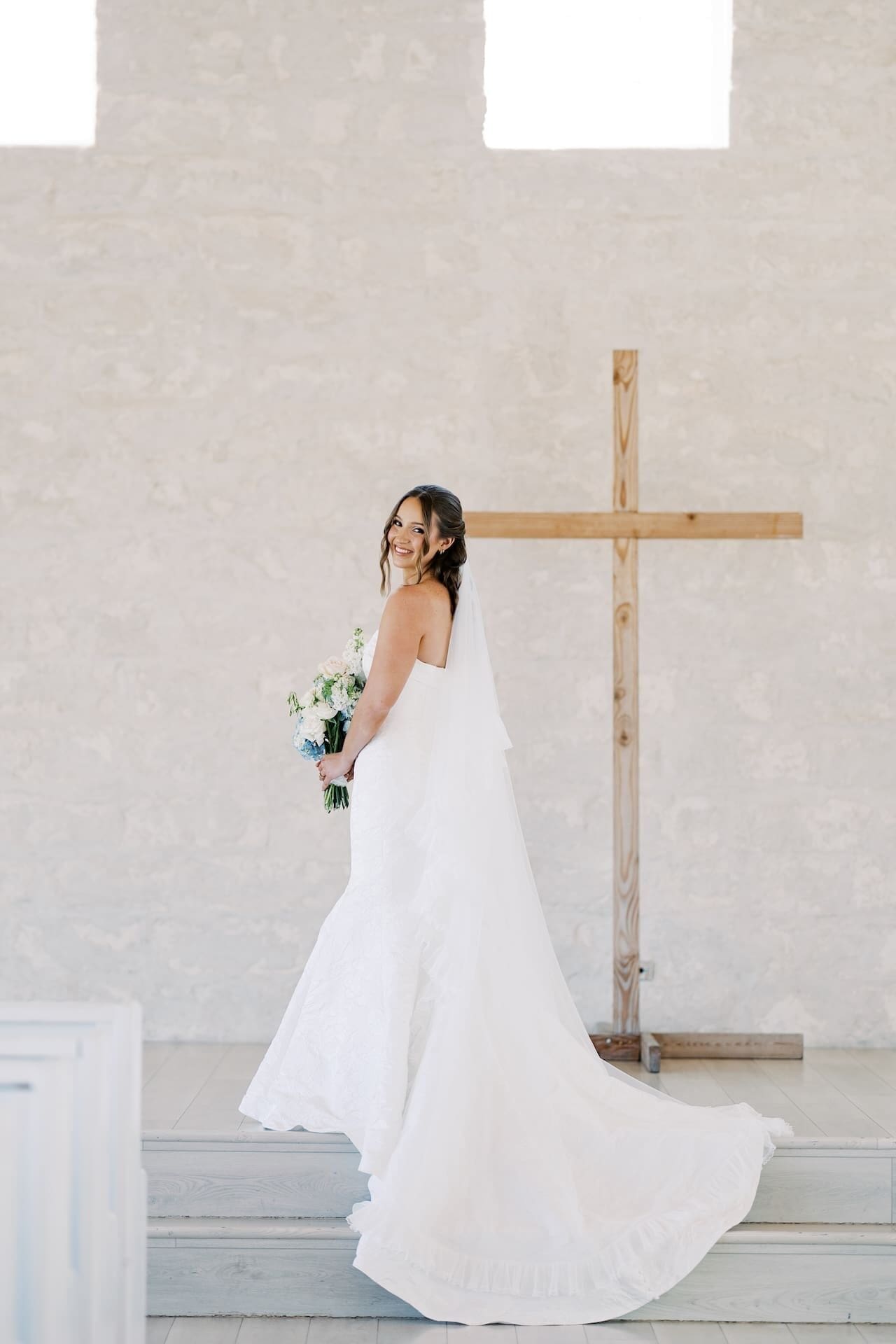 A bride in a strapless mermaid gown glances back with a smile while standing on the chapel altar steps, her cathedral veil and train fanning out beneath a large wooden cross