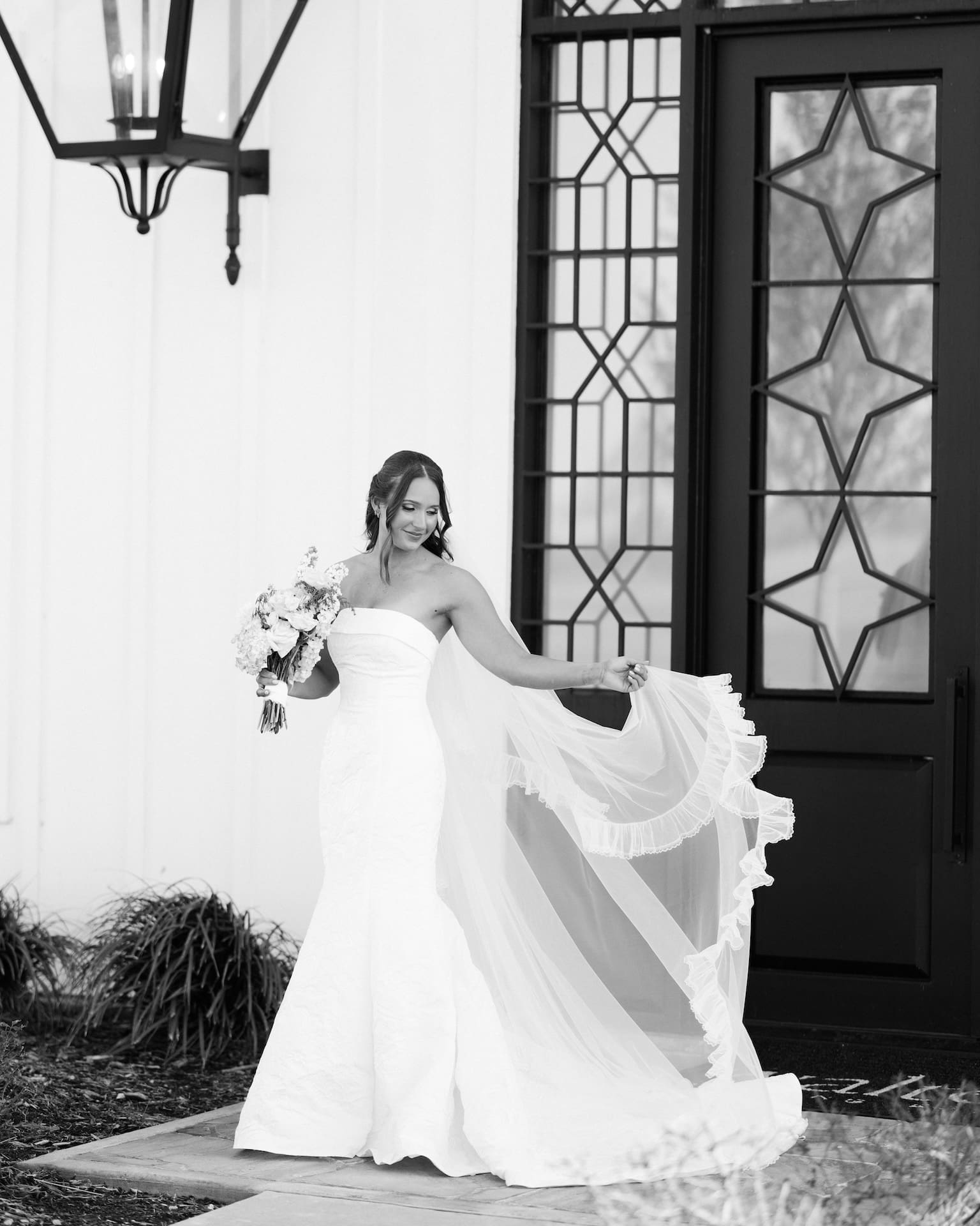 Black and white photo of a bride in a strapless mermaid gown lifting her ruffled veil outside ornate black iron doors of a white barn venue