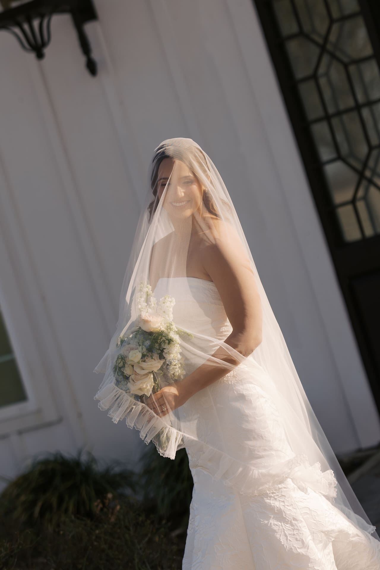 A bride in a strapless lace mermaid gown walks with a smile beneath her flowing veil outside ornate black iron chapel doors in warm golden light